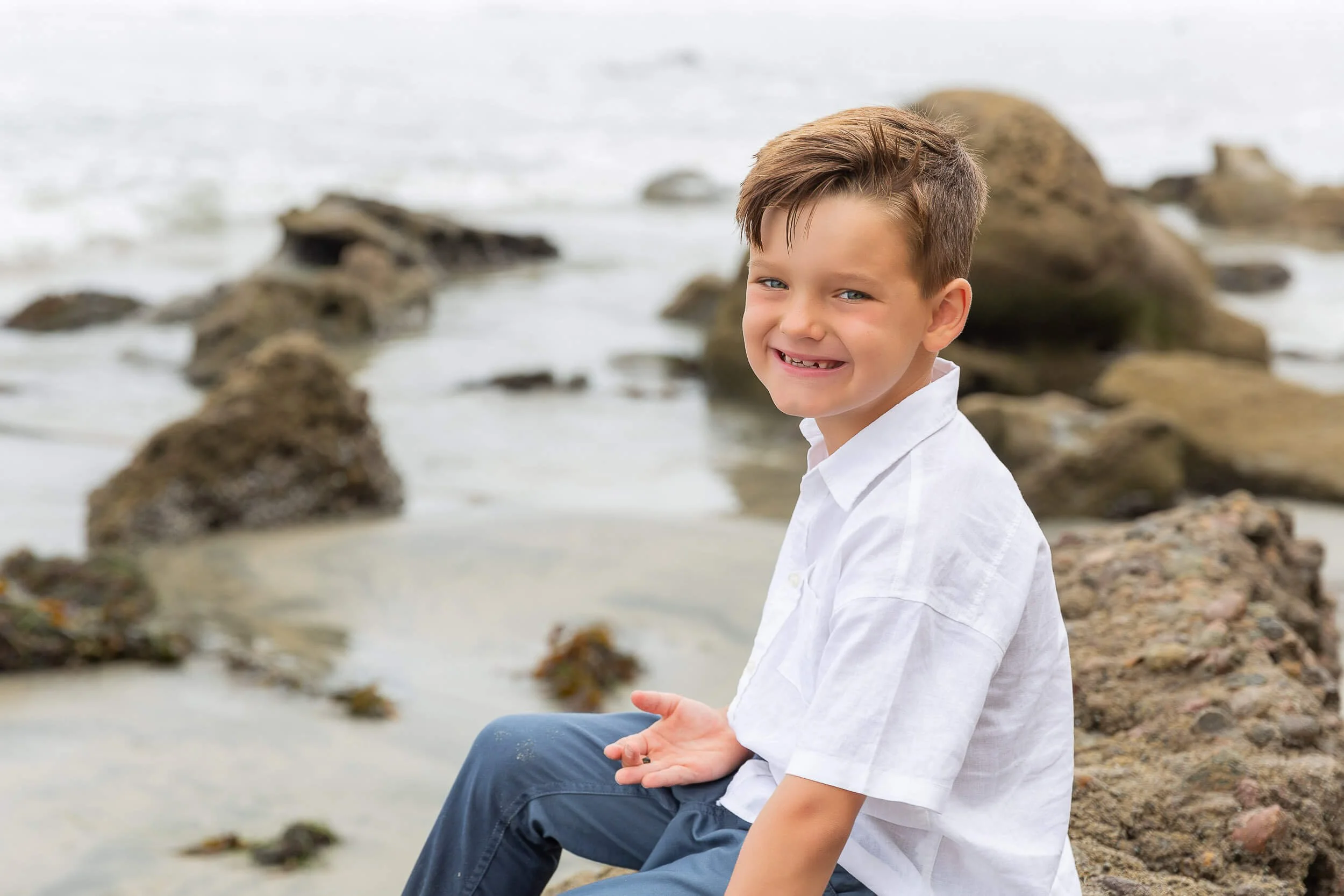 A smiling young boy sitting on rocks at the beach, with ocean waves in the background.