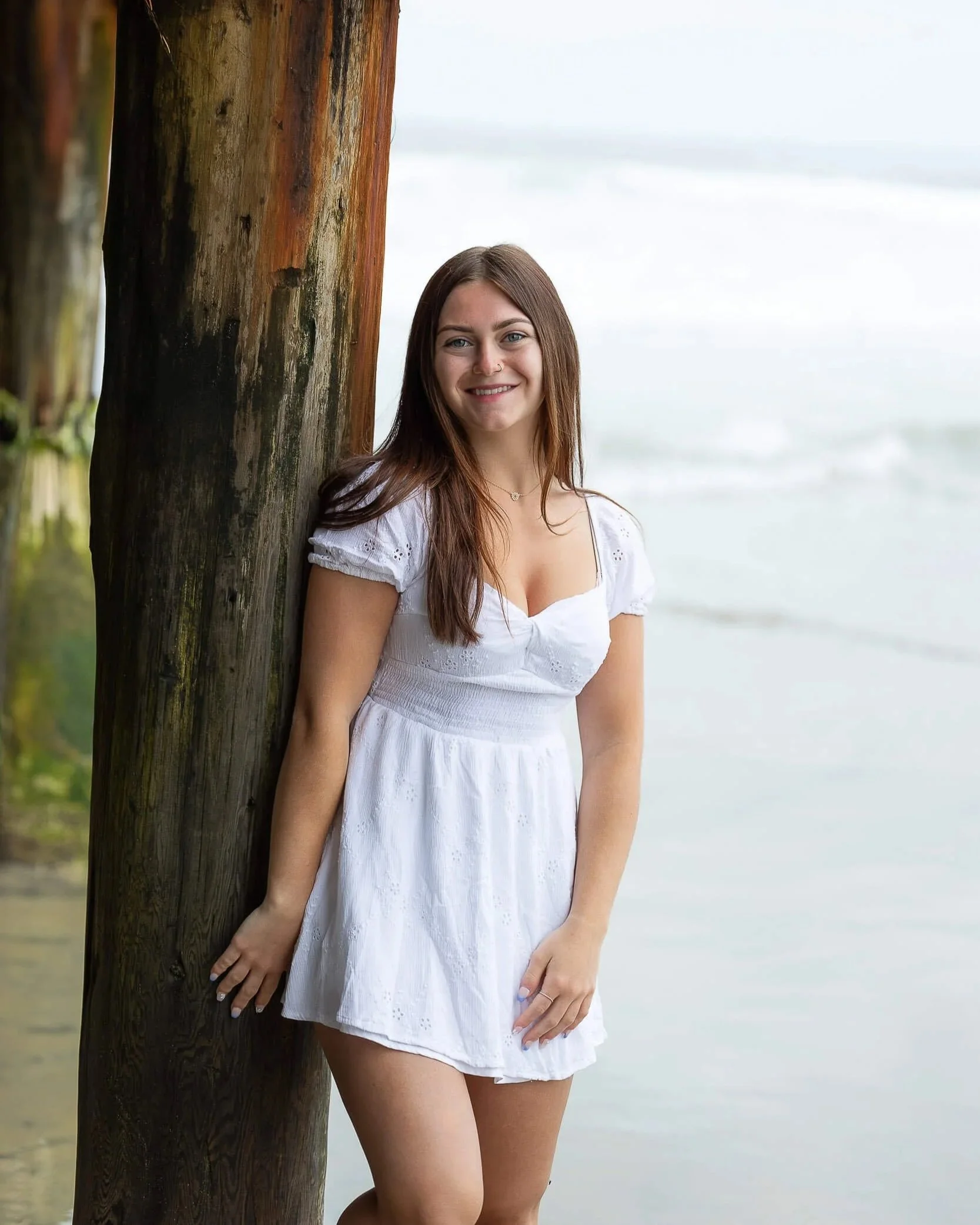 A young woman with long brown hair wearing a white dress, standing and leaning against a wooden pole at the beach with the ocean in the background.