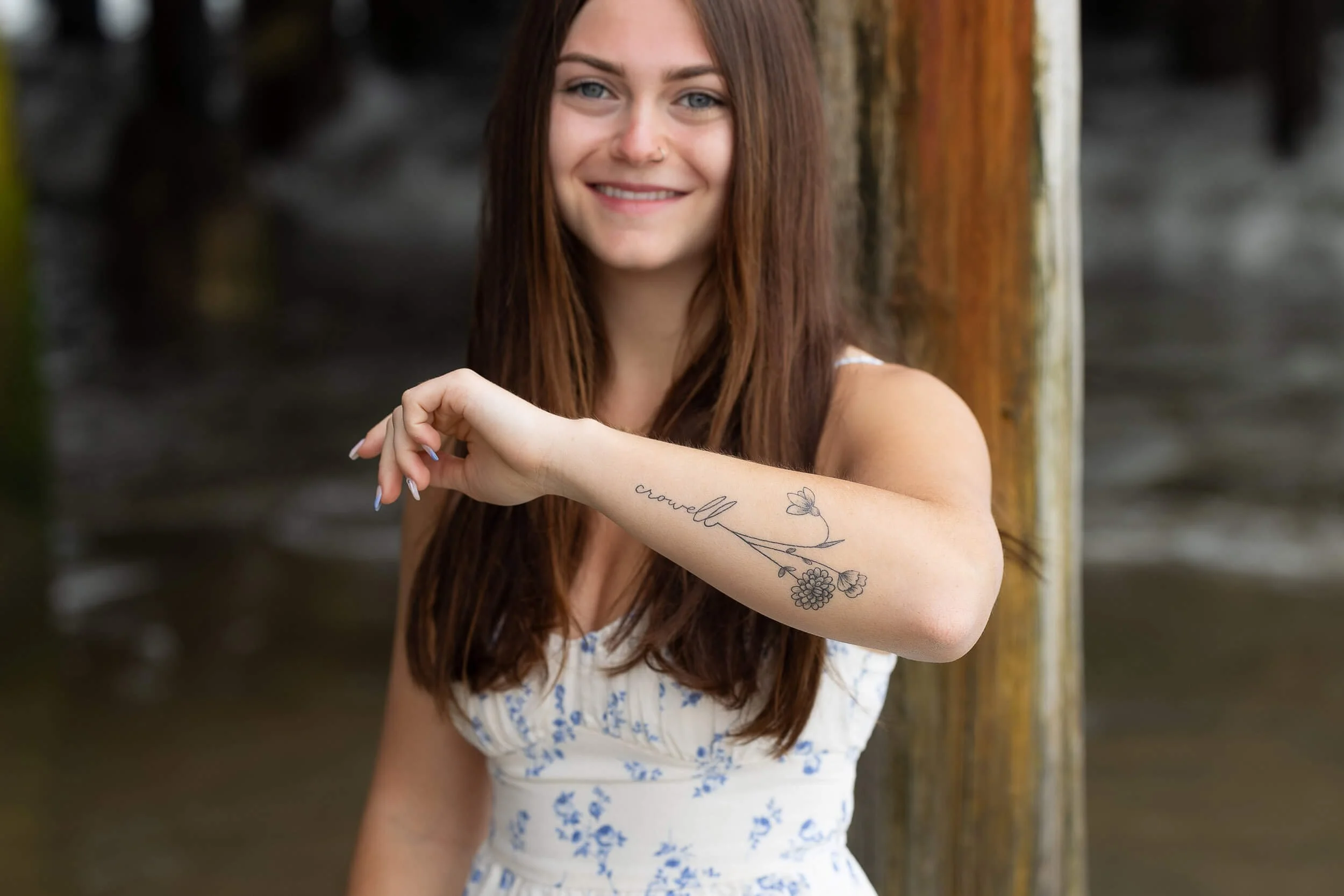 A woman with long brown hair smiling and showing a tattoo with flowers and the word 'crowell' on her forearm, standing outdoors near a wooden post.