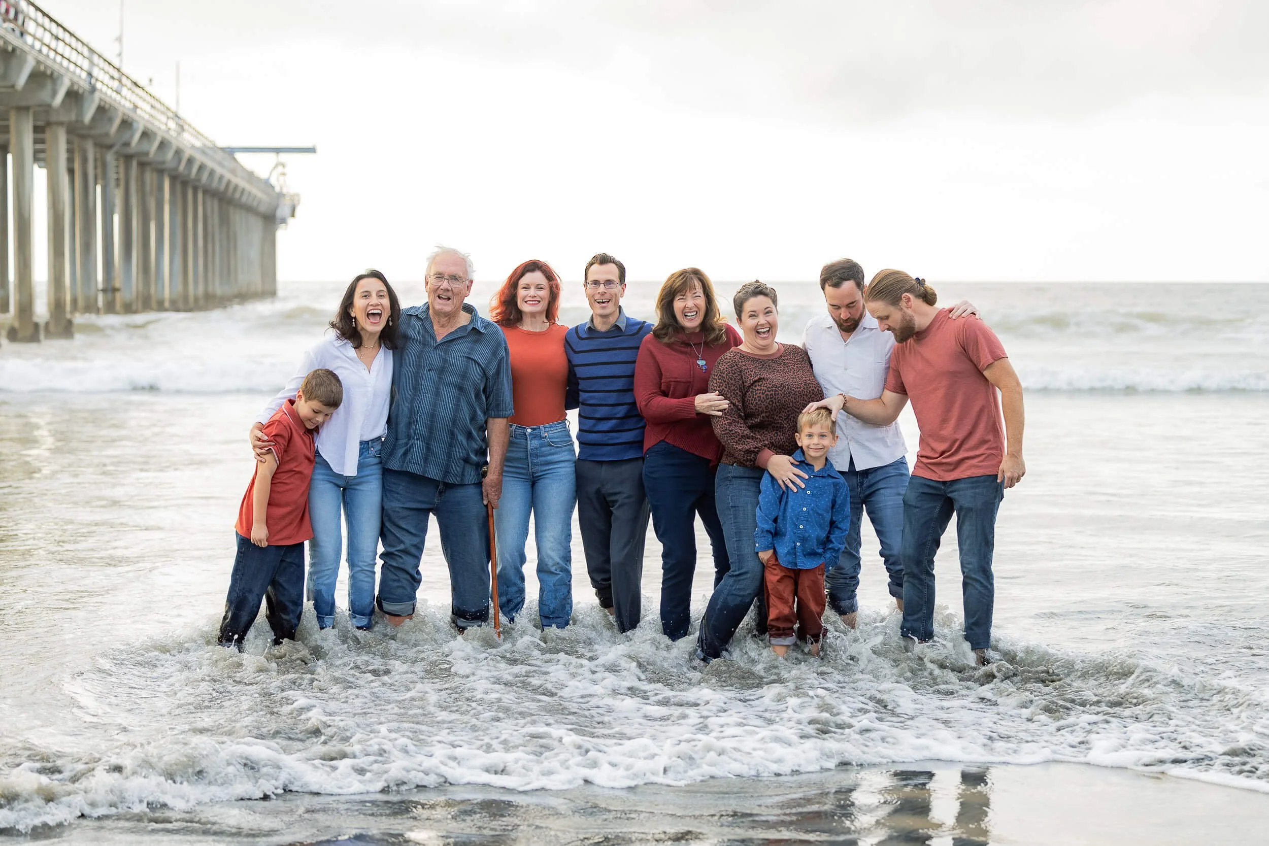 A large group of people, including children, standing in the ocean water near a pier, smiling and enjoying a day at the beach.