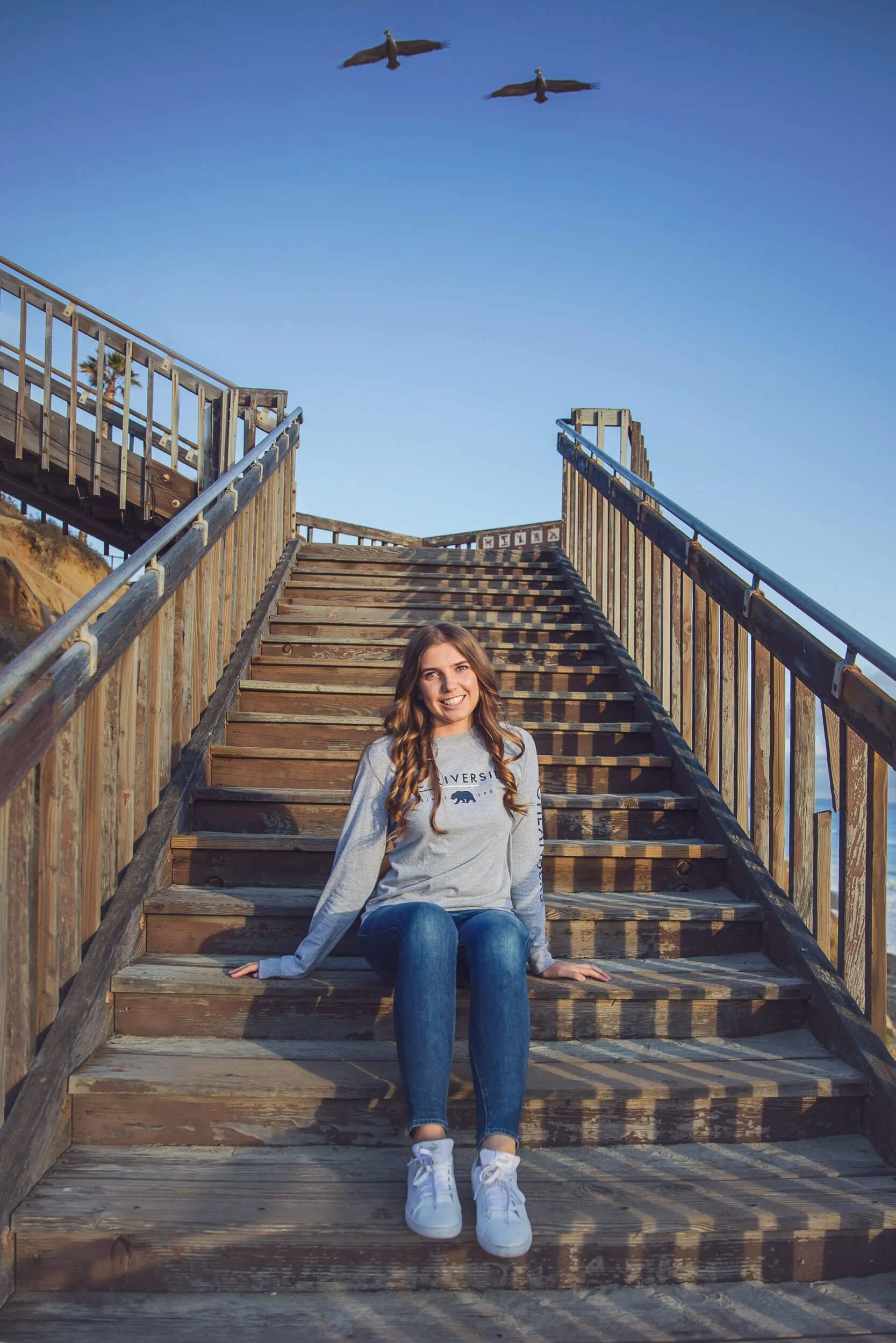 A young woman sitting on wooden stairs outdoors with a blue sky, two seagulls flying overhead, and a palm tree in the background. She is smiling, wearing a gray sweatshirt, jeans, and white sneakers.