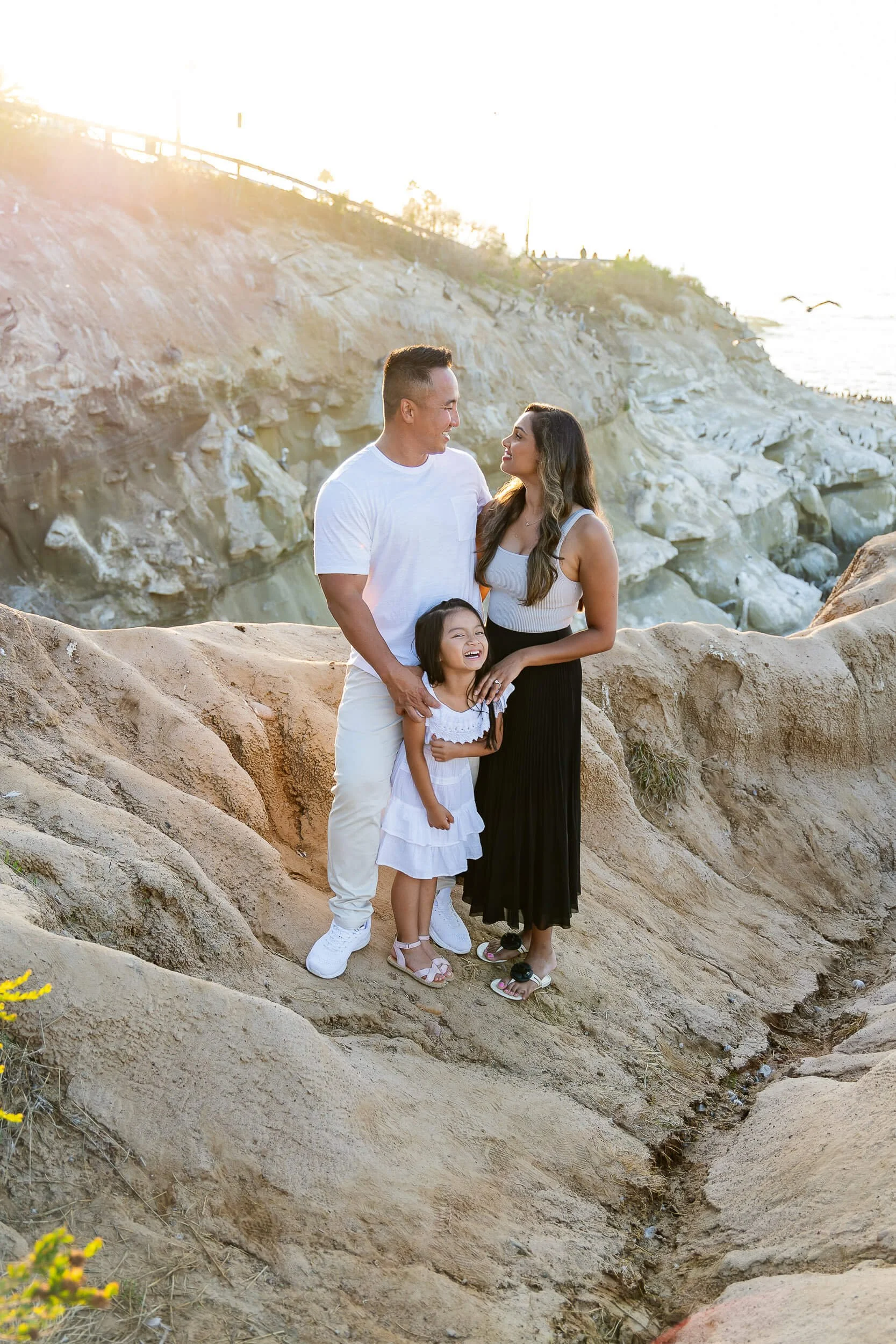 A family of three, a man, woman, and young girl, standing on rocky terrain by the coast during sunset, smiling and looking at each other.