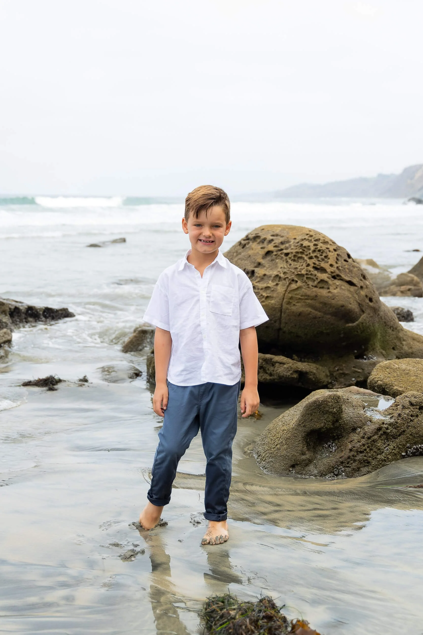 A young boy standing barefoot on a sandy beach with rocks, smiling at the camera, wearing a white shirt and blue pants, with the ocean and overcast sky in the background.