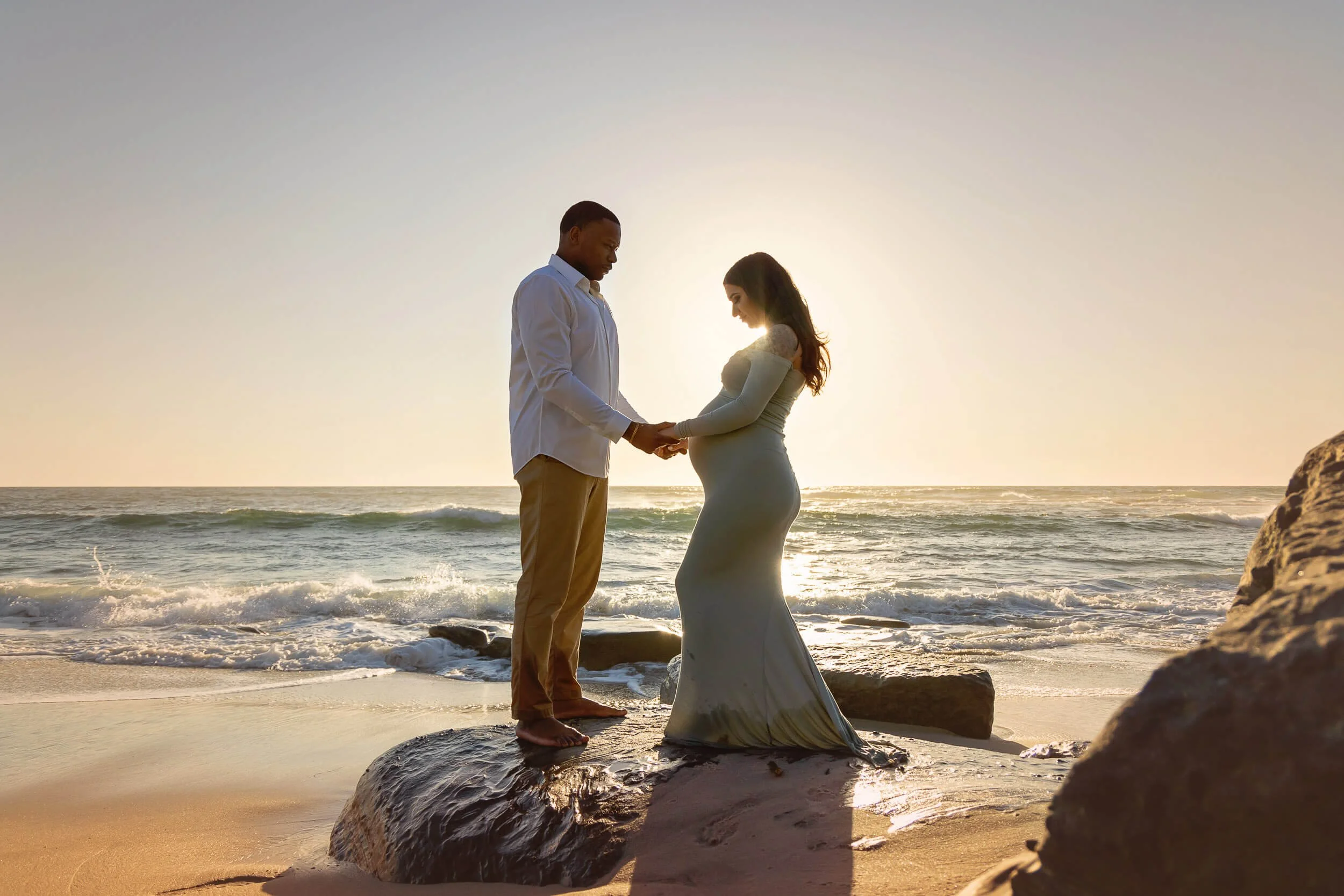 A couple holding hands on a rock at the beach during sunset, with the pregnant woman in a long dress and the man in a white shirt and pants.