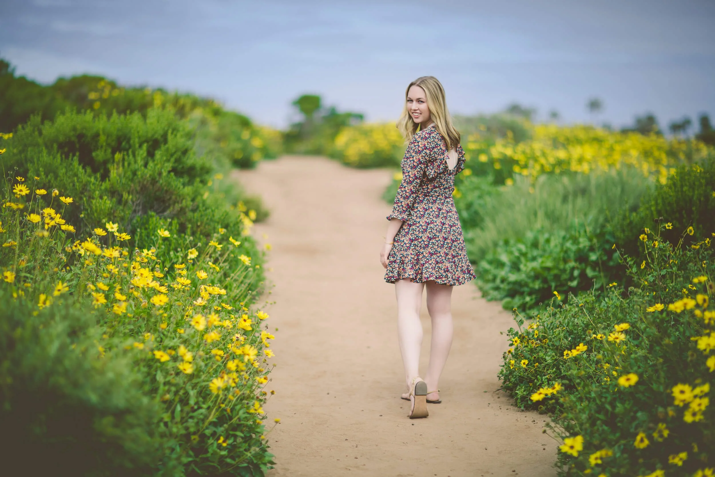 A woman in a floral dress walking on a dirt path surrounded by yellow wildflowers.