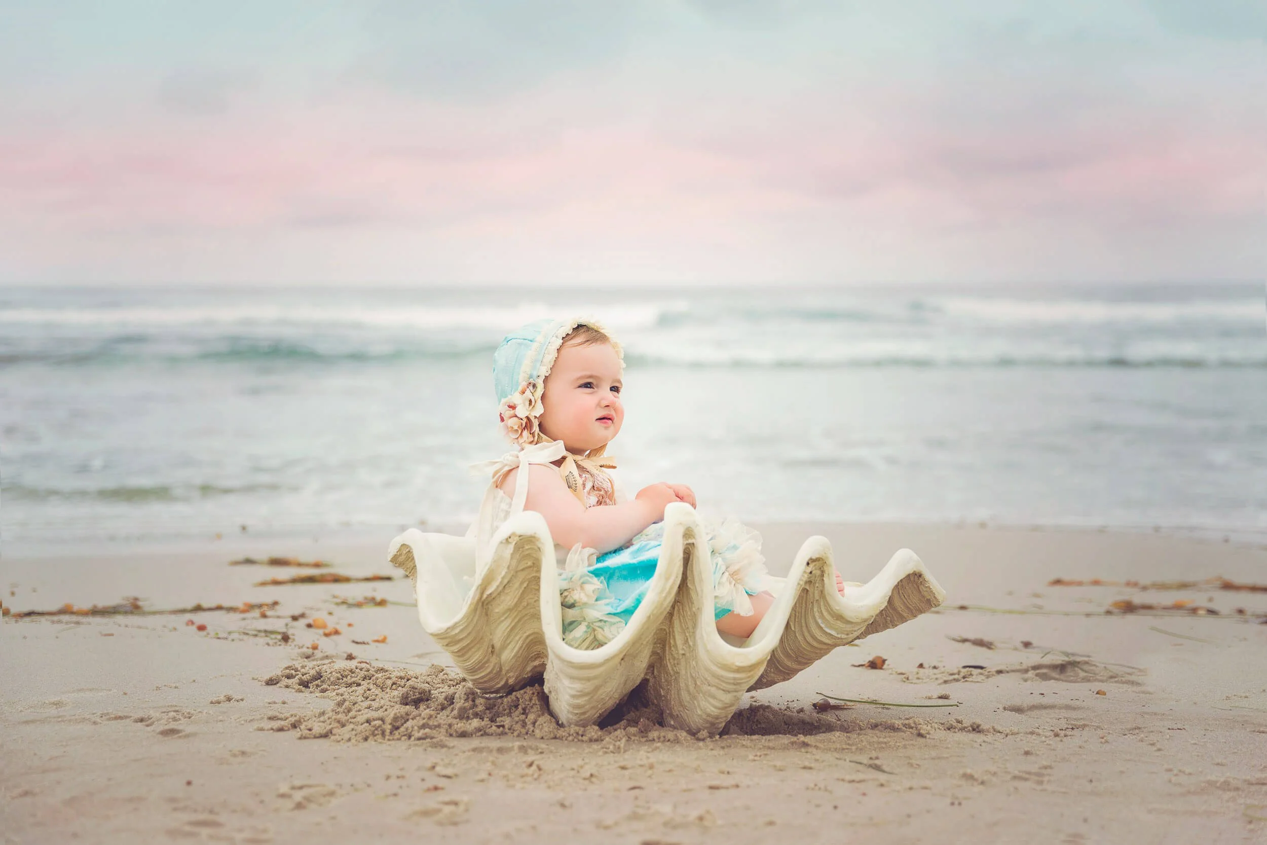 A young child sitting inside a large seashell on the sandy beach, wearing a bonnet and light-colored dress, with the ocean and sky in the background.