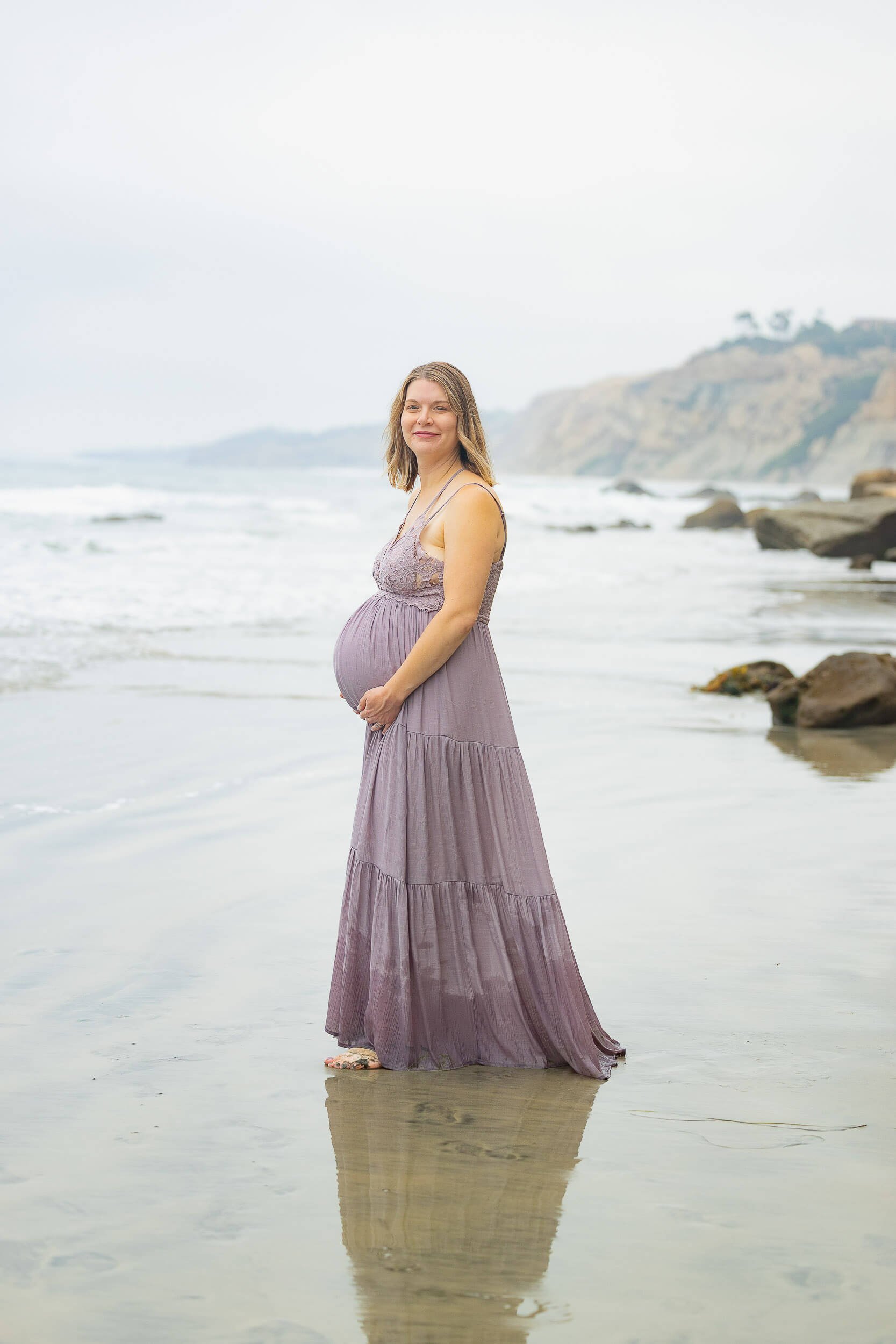 A pregnant woman standing on the beach in a purple dress, smiling, with the ocean and rocky cliffs in the background.
