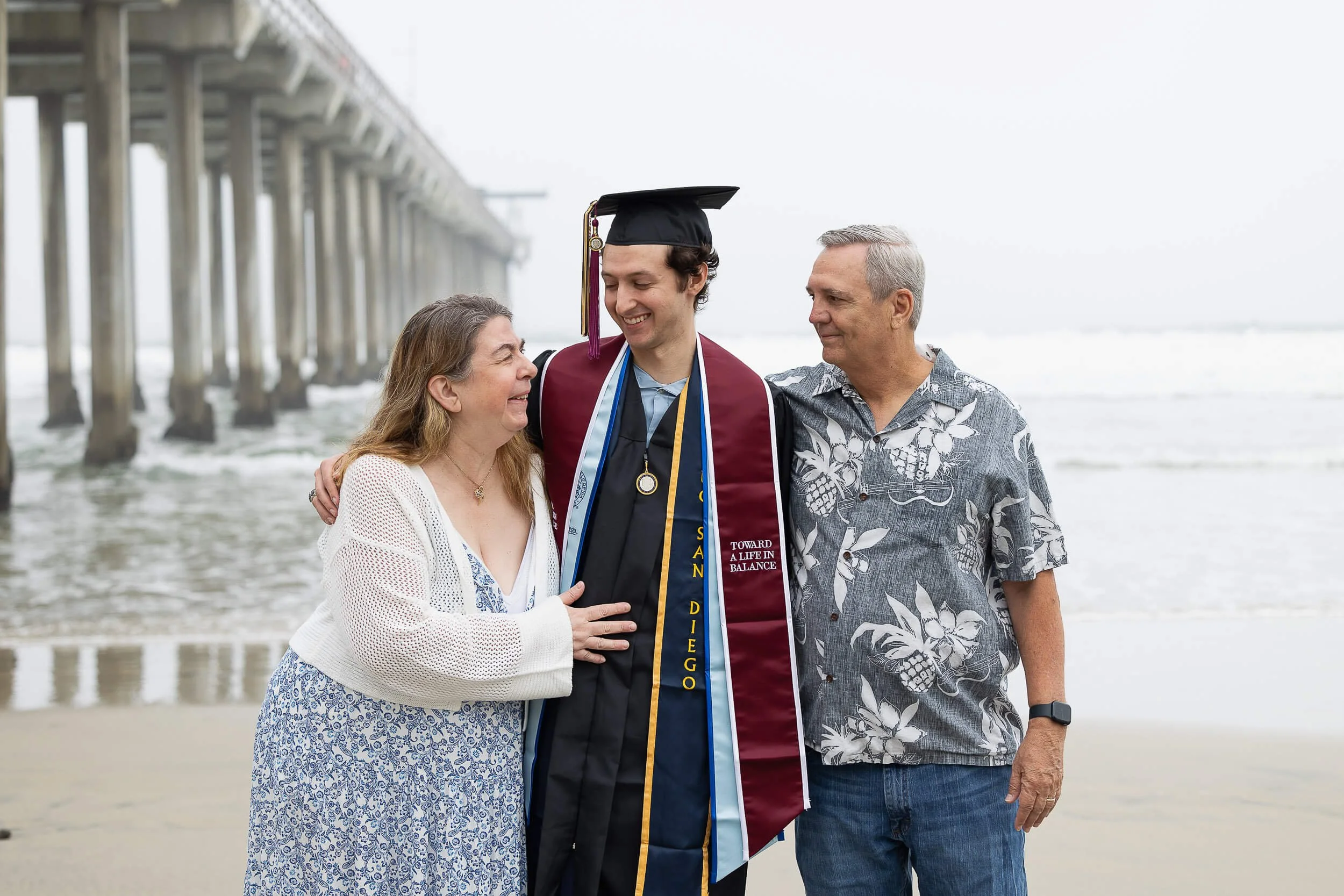 A young man in a graduation cap and gown on the beach, flanked by his parents, all smiling and looking at each other.