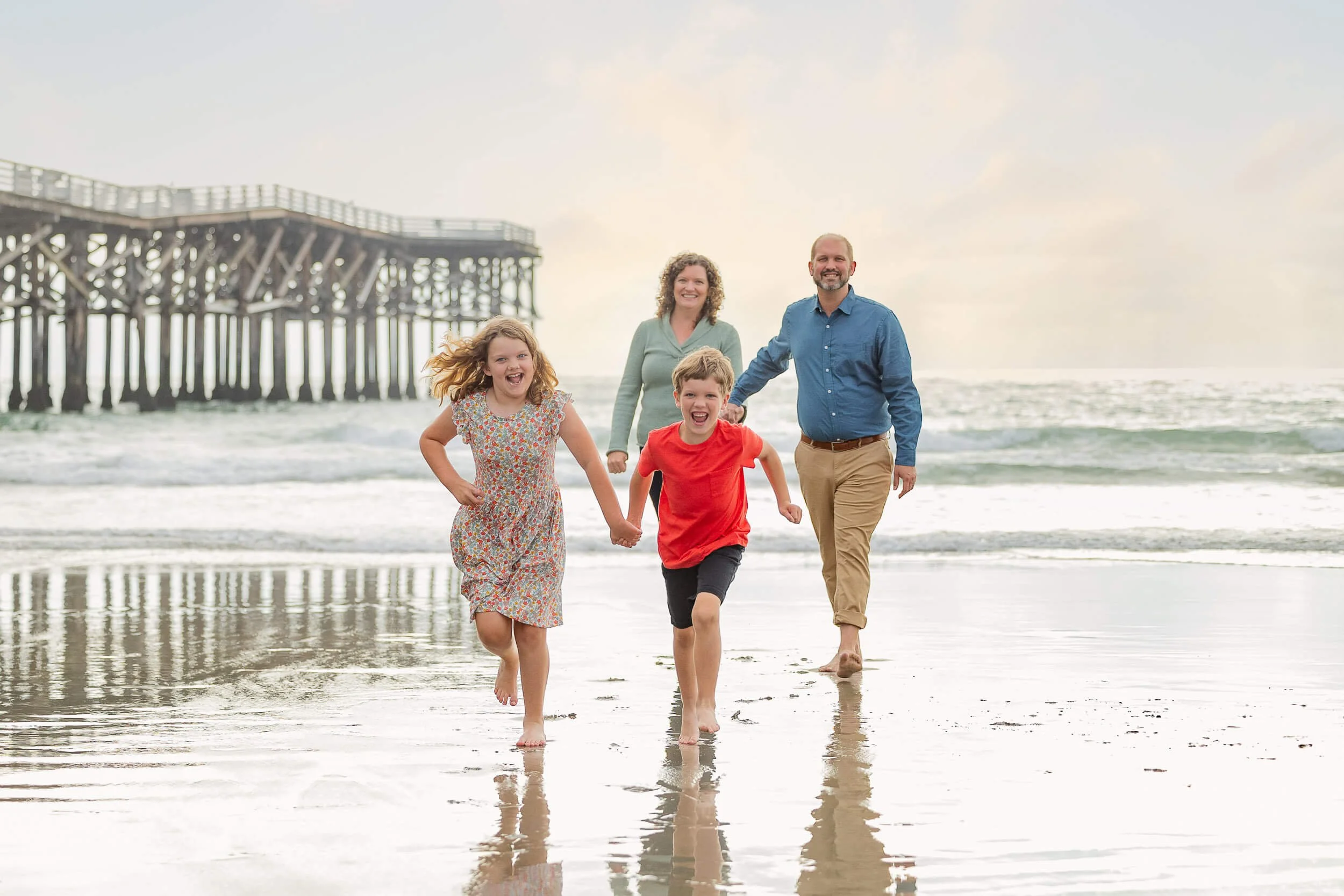 Family of four running on the beach, holding hands, with a pier in the background and the ocean in view.