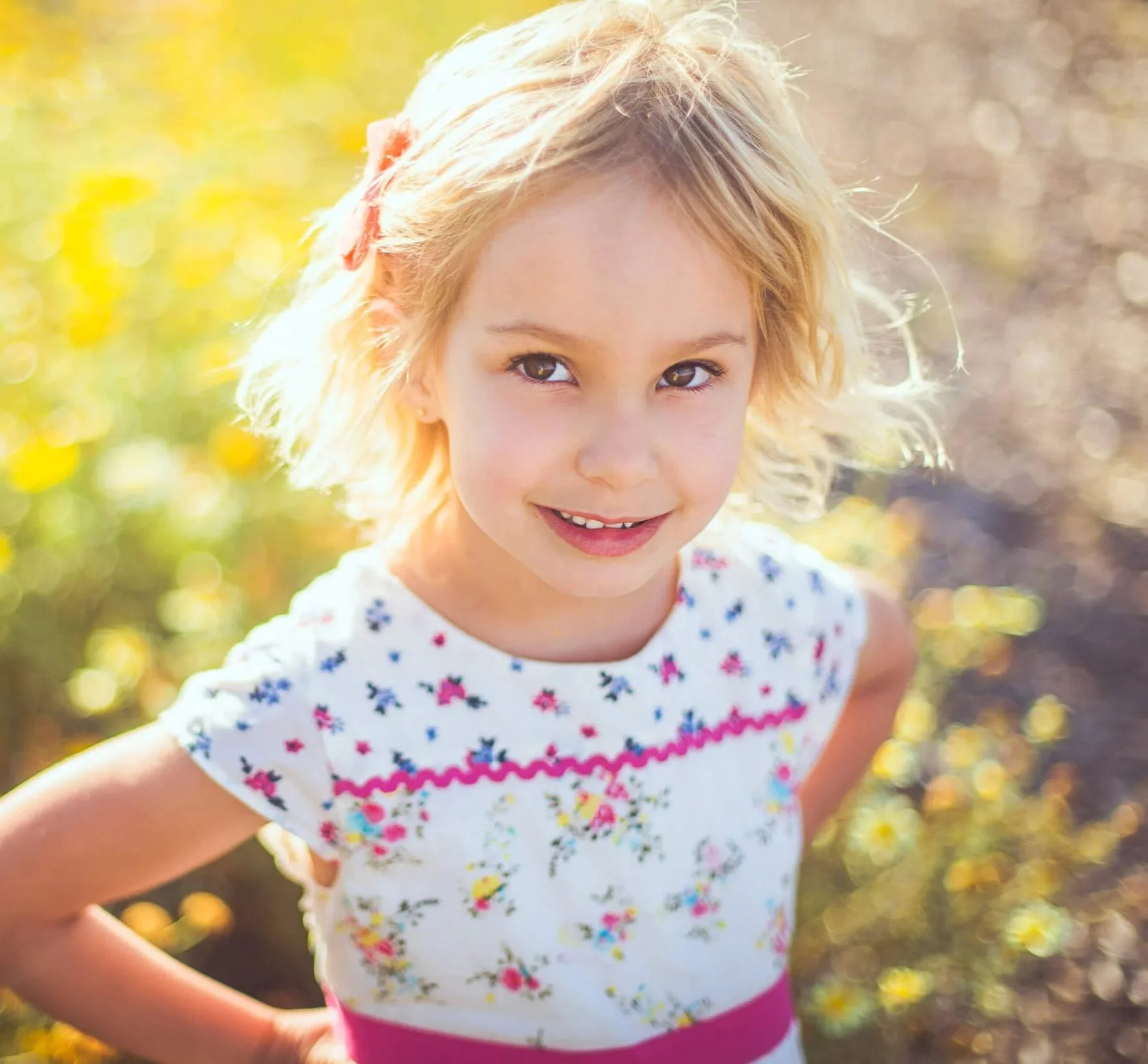A young girl with blonde hair and a flower hair clip outdoors during daytime, smiling at the camera, wearing a white dress with colorful floral patterns and pink accents.