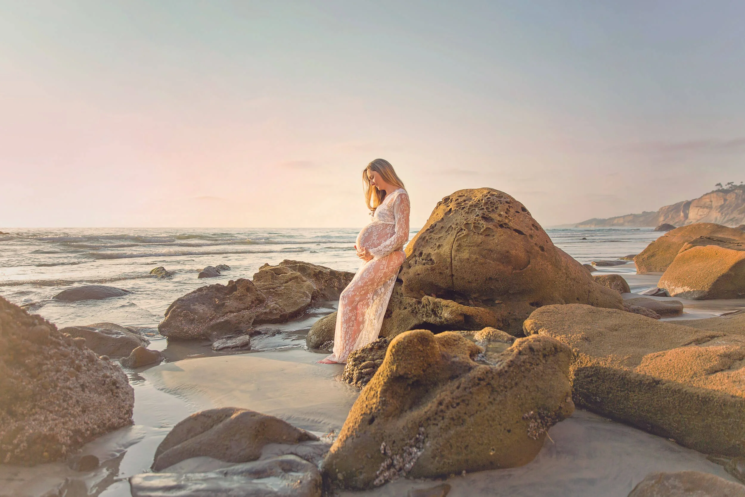 A pregnant woman wearing a white lace dress stands on rocks by the beach during sunset, caressing her belly.