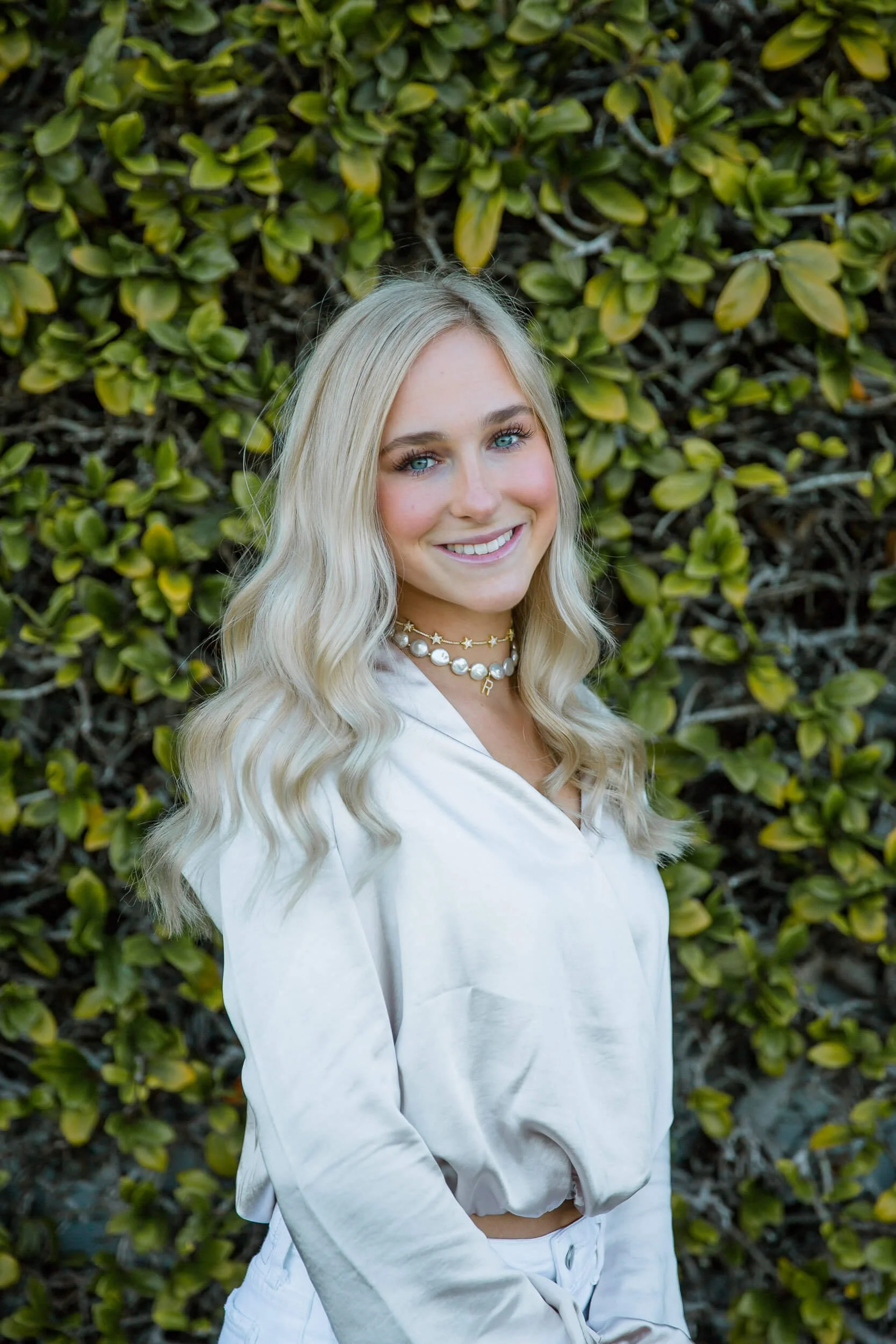 A young woman with long blonde hair smiling outdoors against a leafy green background, wearing a white satin blouse and layered pearl necklaces.
