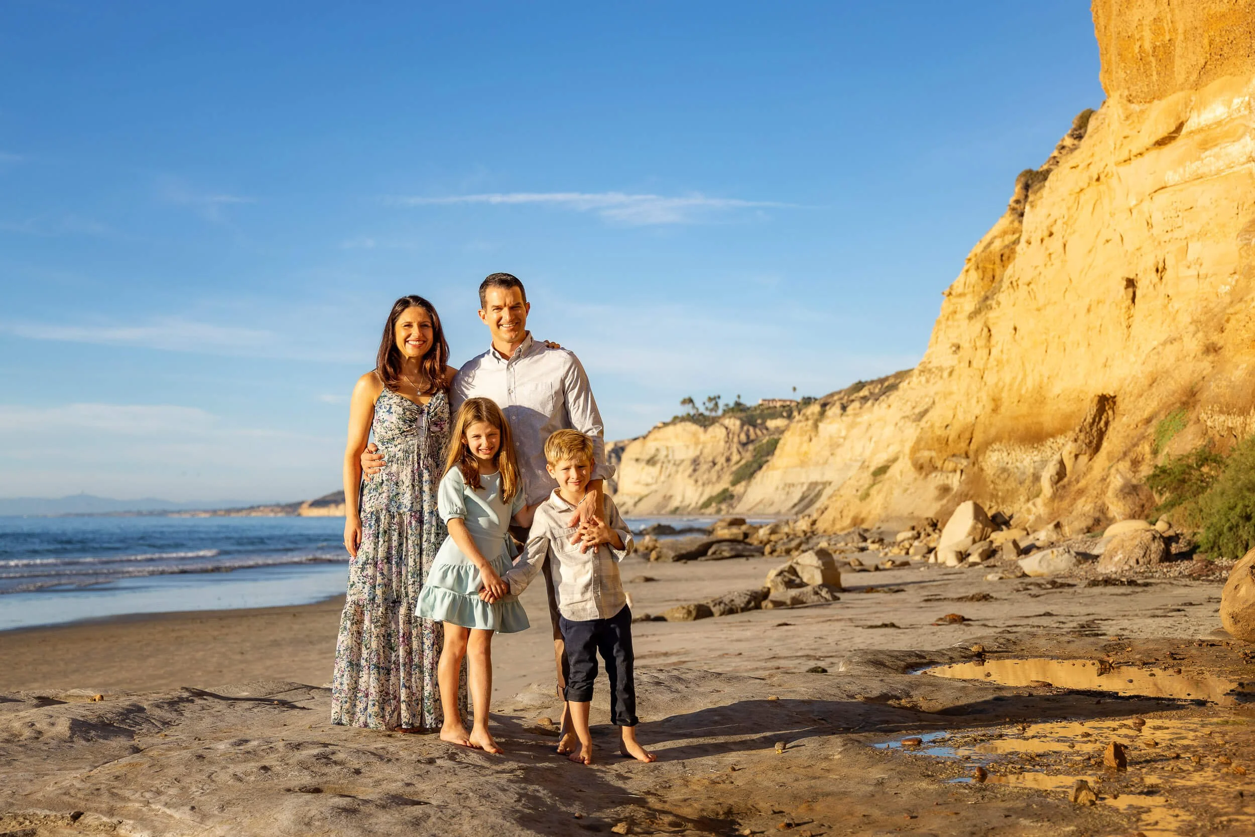 A family of five standing on a beach with cliffs in the background during sunset, smiling at the camera.