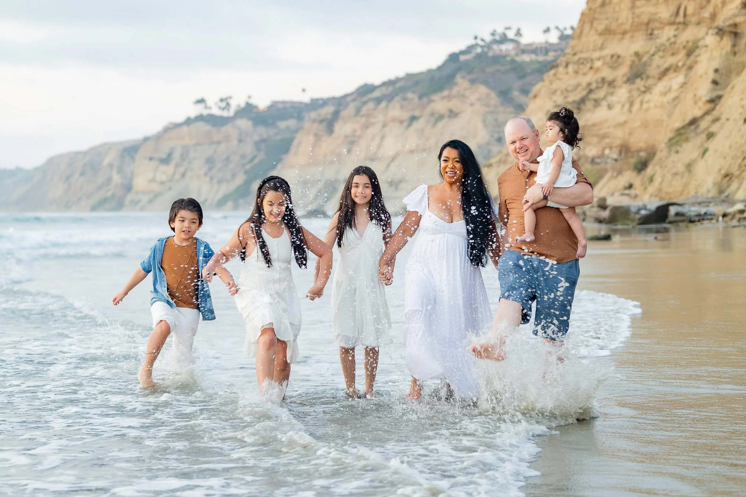 A multiethnic group of six people, including children and adults, joyfully walking in the ocean waves on a beach with cliffs in the background.