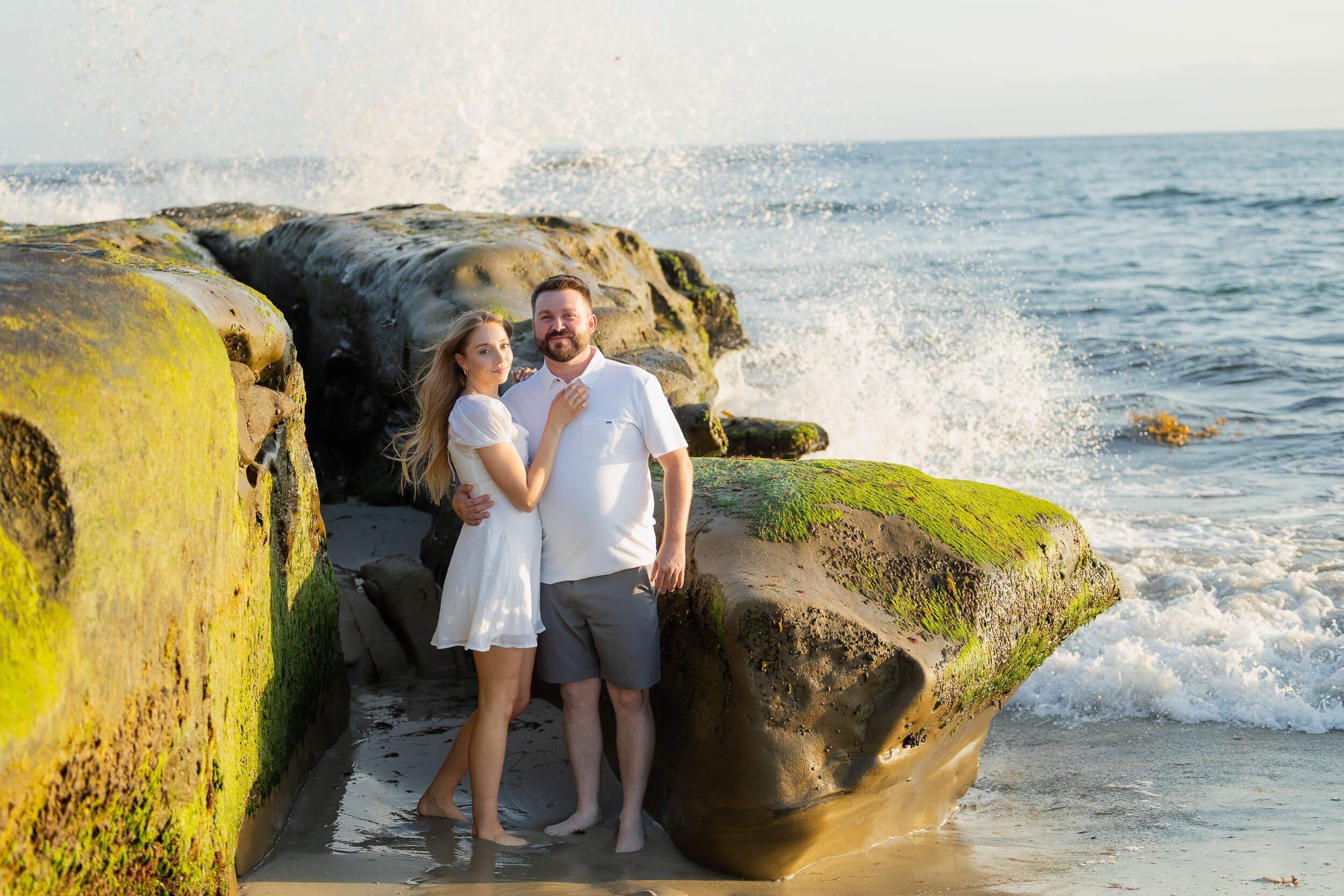 A young couple standing on the beach with large moss-covered rocks and the ocean in the background