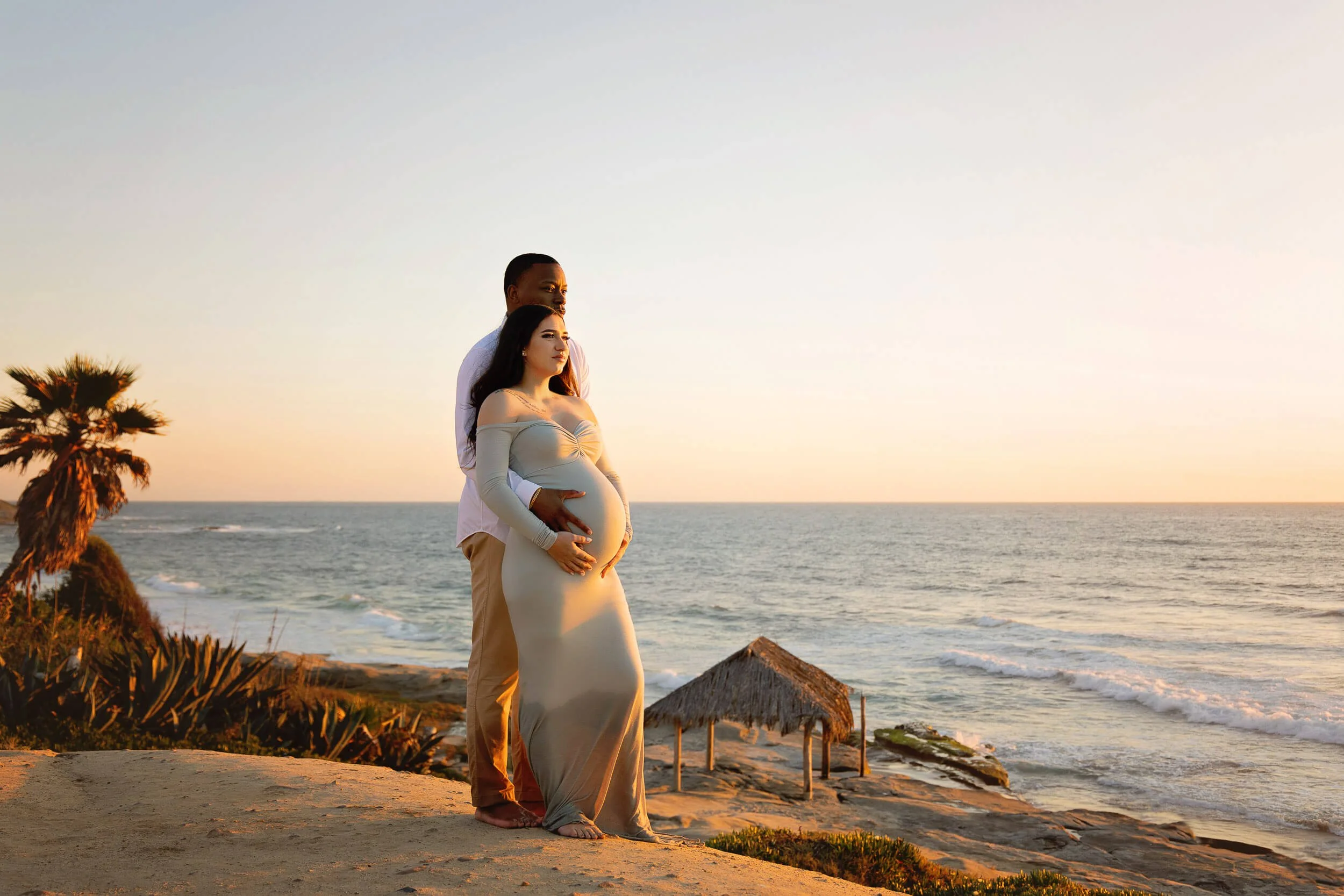 A pregnant woman and a man standing on a beach during sunset, with the woman in a long off-shoulder dress and the man embracing her from behind.