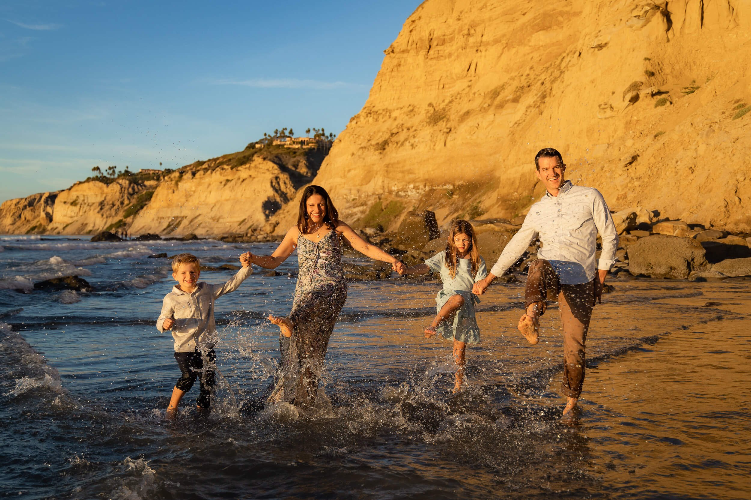 A family of four playing and jumping in the ocean at sunset, with cliffs and a house in the background.