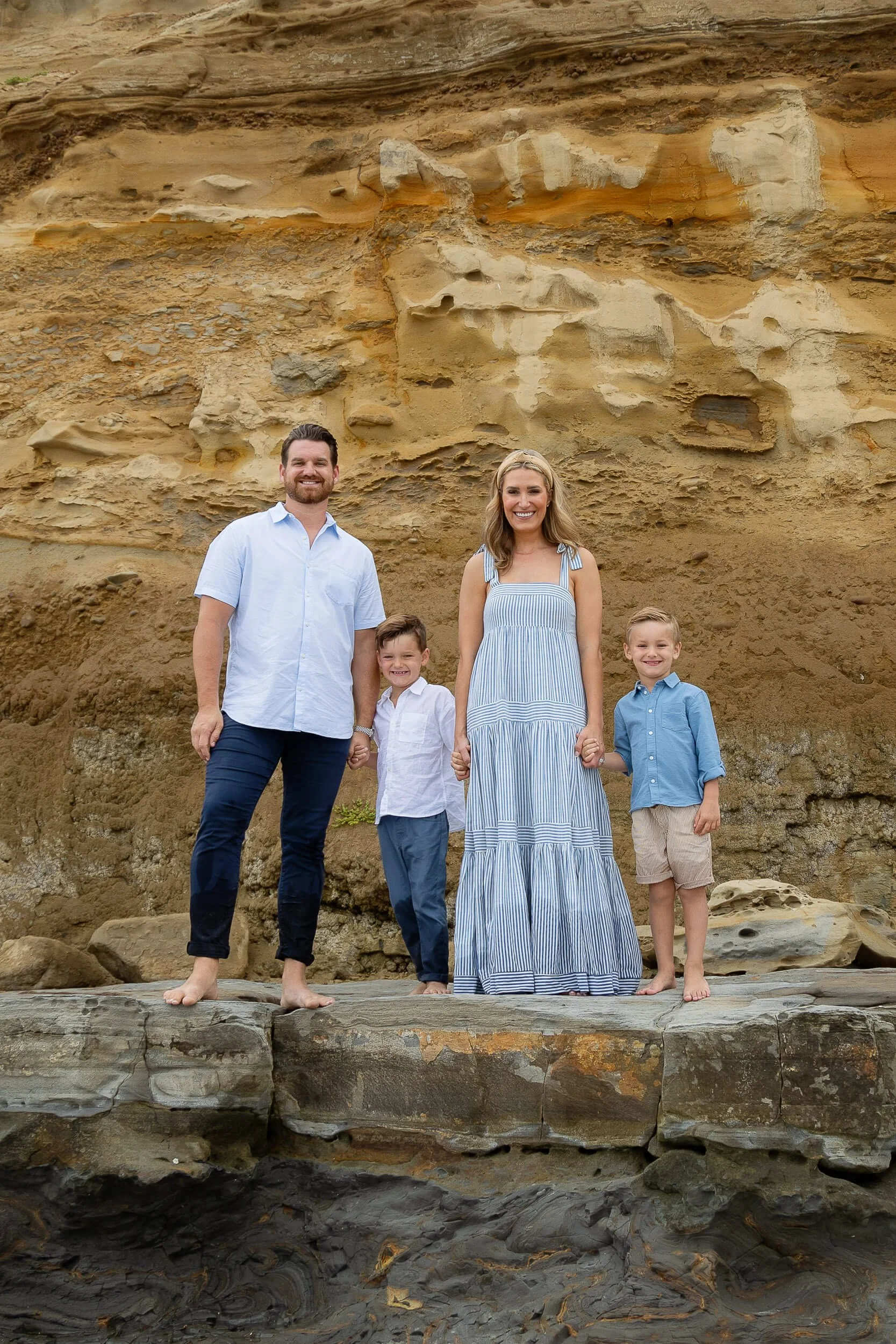 Family of four standing barefoot on rocks at the beach, holding hands, with a sandy cliff in the background.