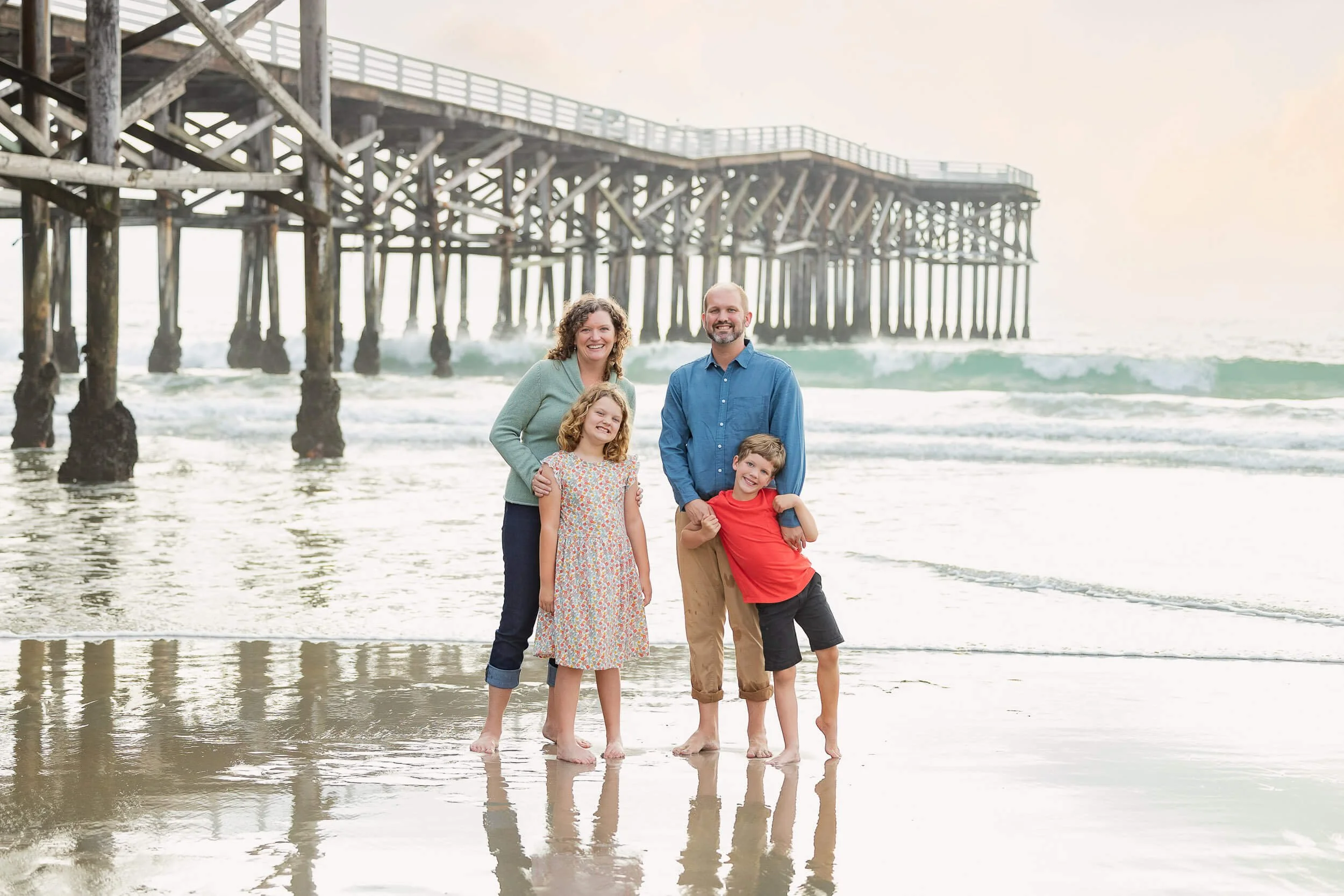 A family of four standing on a beach near a wooden pier, smiling at the camera, with the ocean and waves in the background.