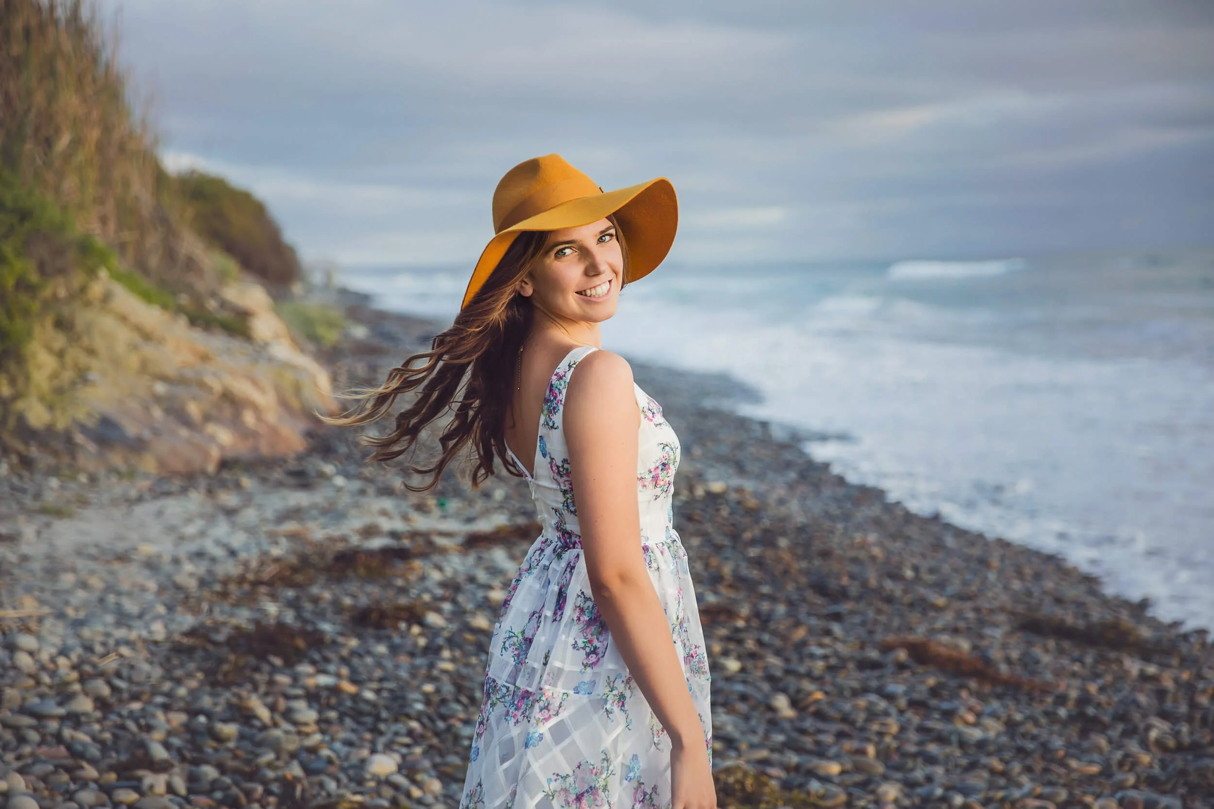 Smiling young woman in a white floral dress and wide-brimmed hat standing on a rocky beach facing the ocean with cliffs in the background.