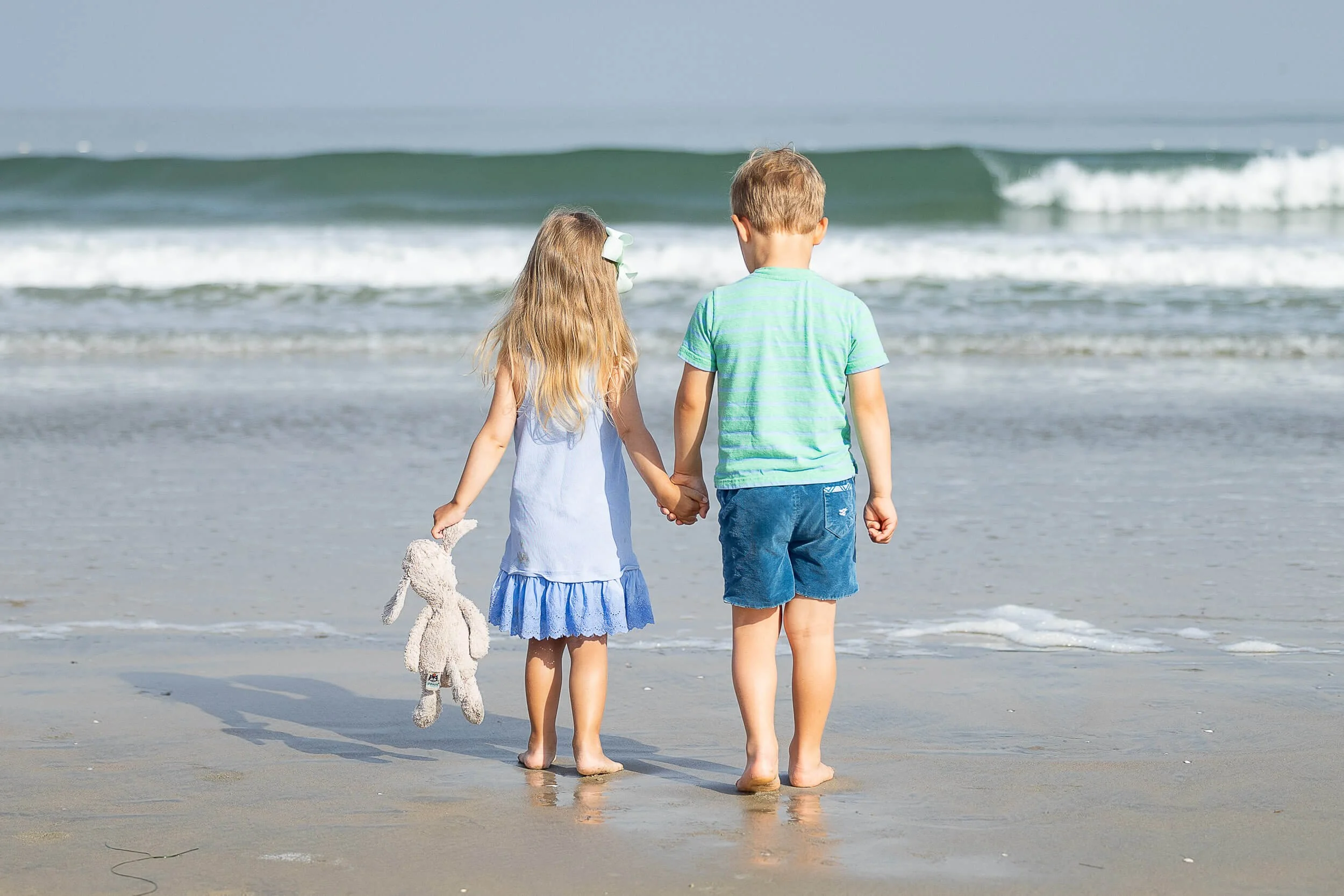 Two children, a girl and a boy, holding hands and walking on the beach near the water, with the girl holding a stuffed animal.