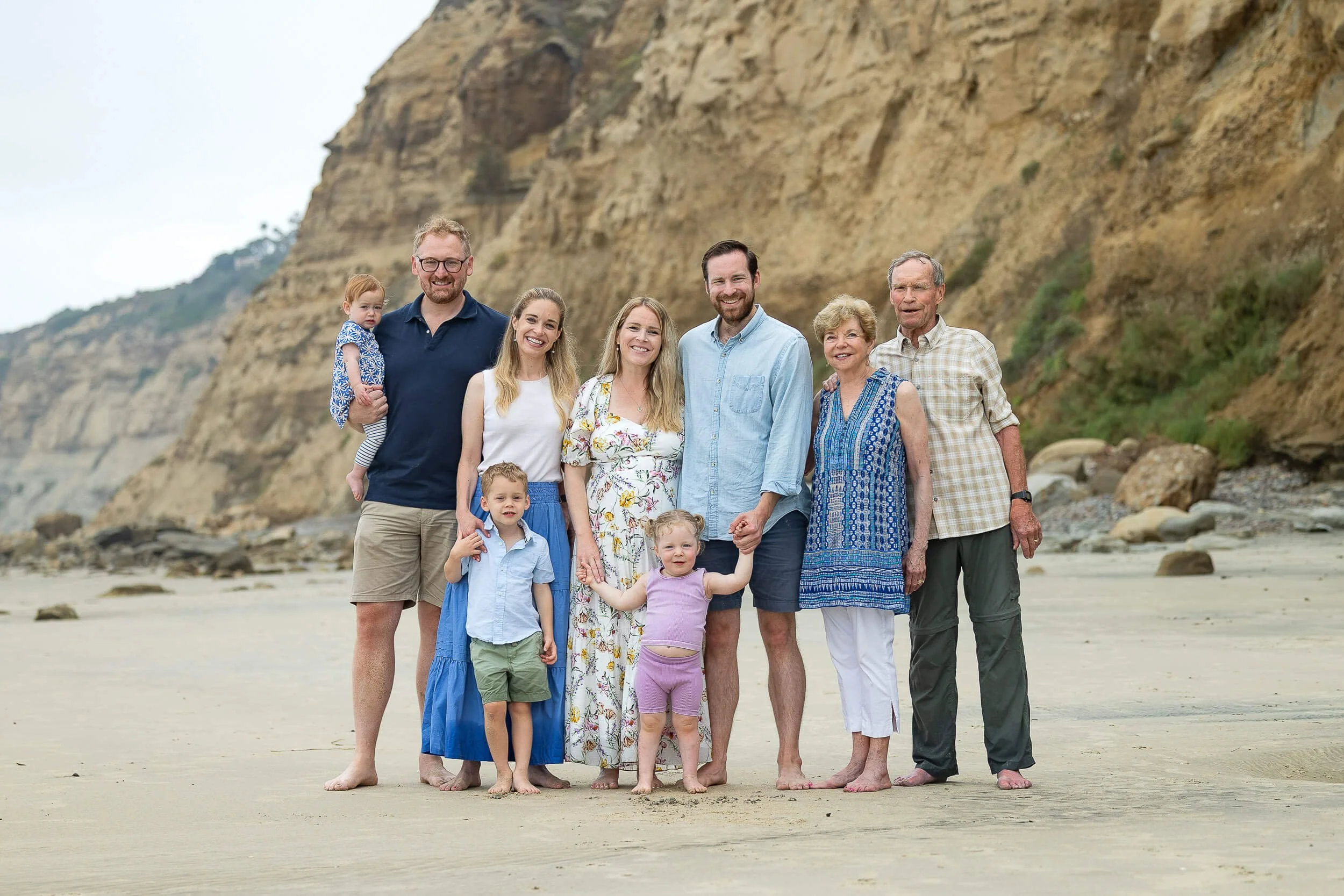 A multi-generational family standing on a sandy beach with cliffs in the background, smiling at the camera.