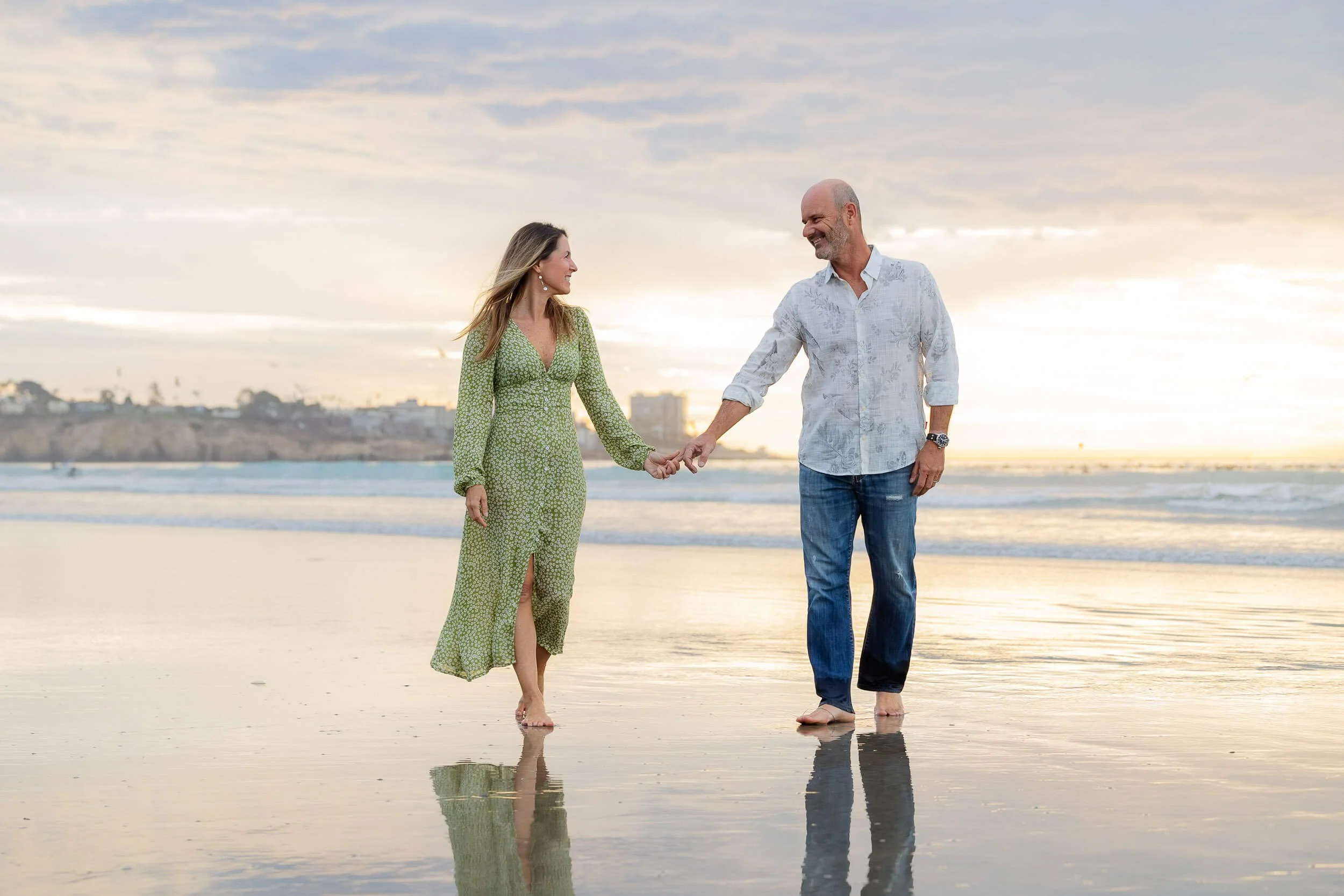 A couple holding hands and walking on the beach at sunset, smiling and looking at each other.