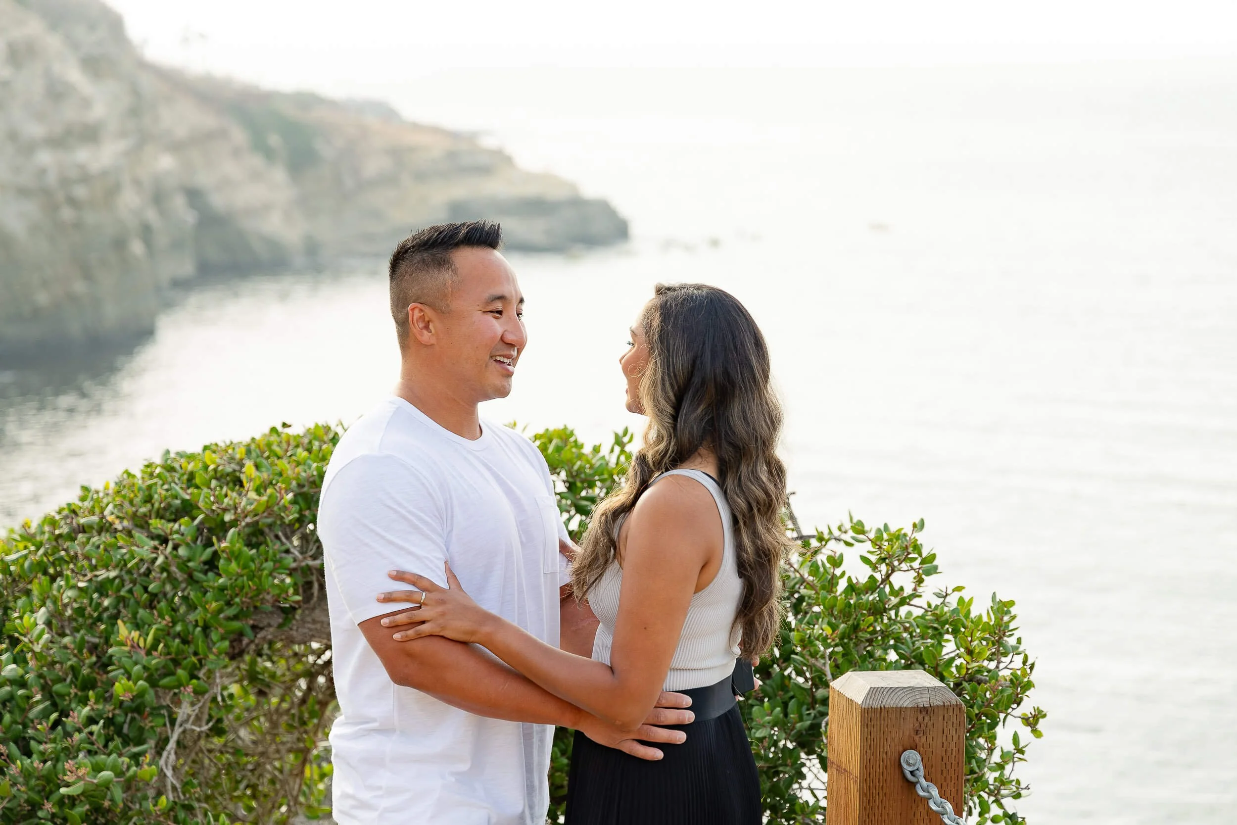 A couple stands close to each other on a scenic coastline, smiling at each other, with lush green bushes and the ocean in the background.