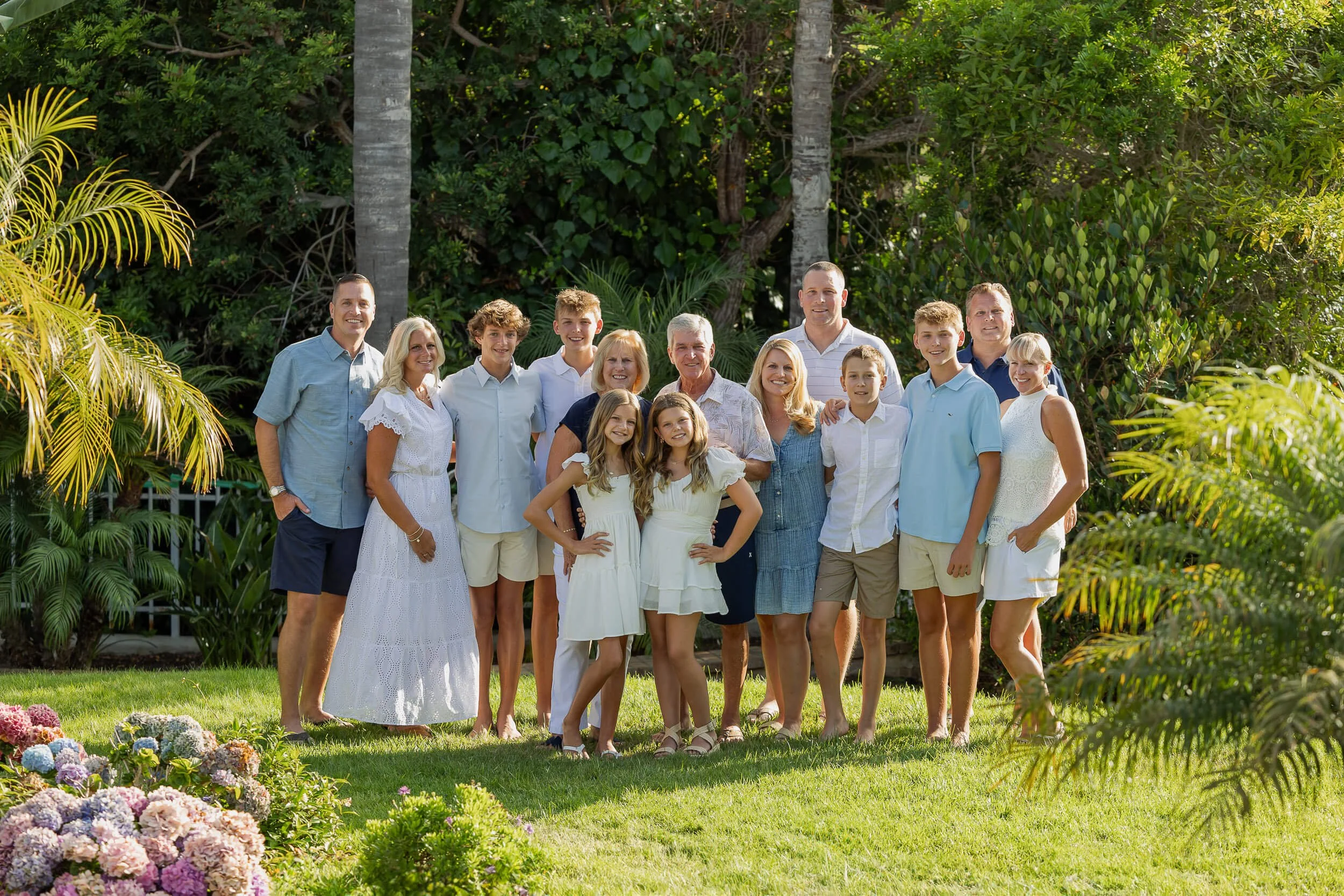 Family group photo outdoors in a lush garden with green trees and plants, including colorful flowers in the foreground. The group consists of adults and children dressed in casual summer clothes, smiling at the camera.