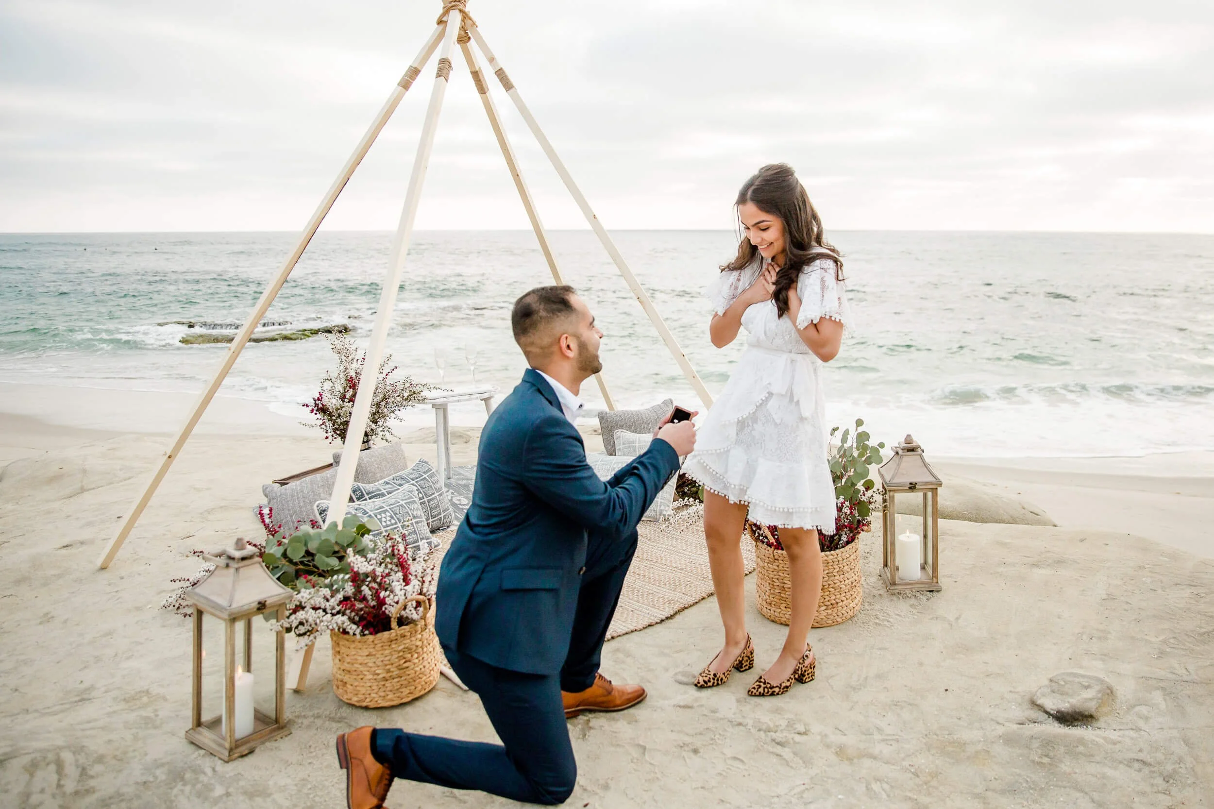 A man proposes to a woman on the beach during sunset, with the ocean and cloudy sky in the background. The man is kneeling holding a ring, while the woman looks surprised and emotional. They are surrounded by decorative lanterns, baskets with flowers, and pillows under a wooden teepee structure.