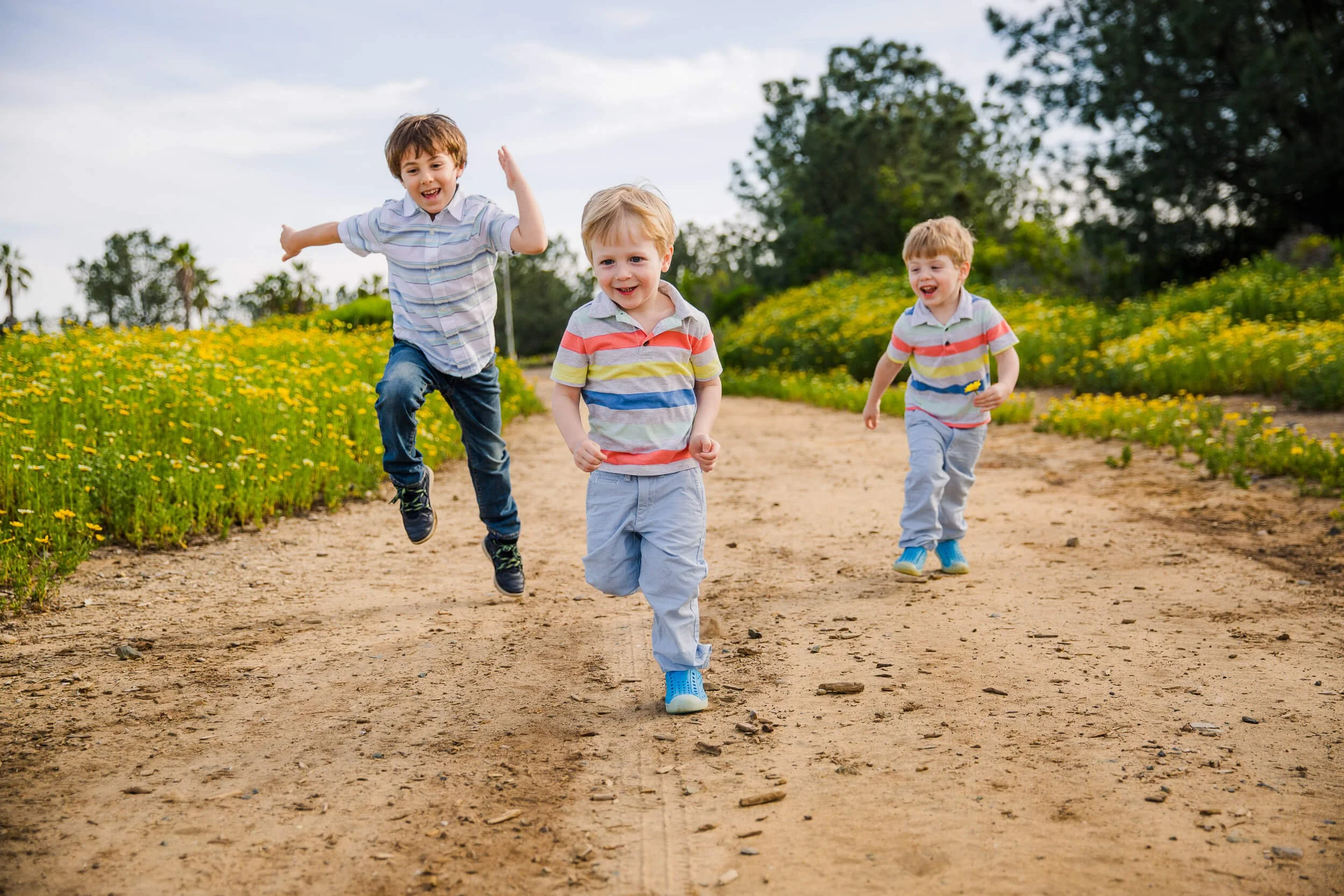 Three young boys running and playing on a dirt path surrounded by yellow flowers and greenery on a sunny day.