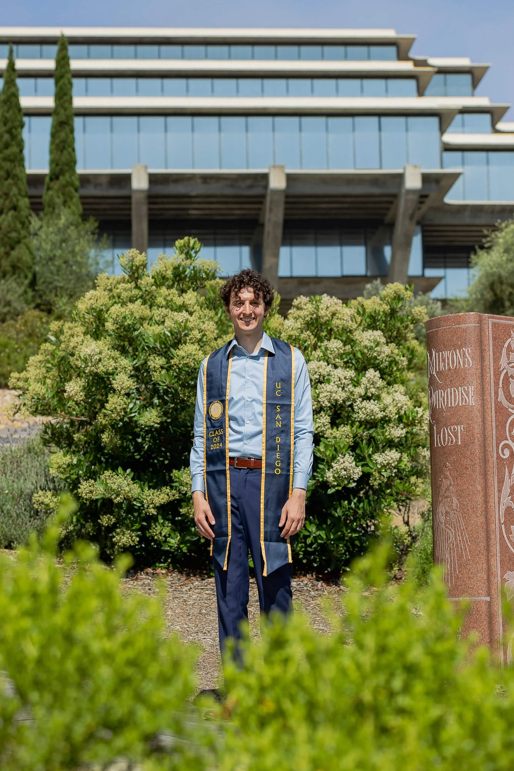 A young man in a blue shirt and slacks stands outdoors, wearing a graduation stole that reads 'Class of 2024' and 'UC San Diego,' with a modern building and green bushes in the background.