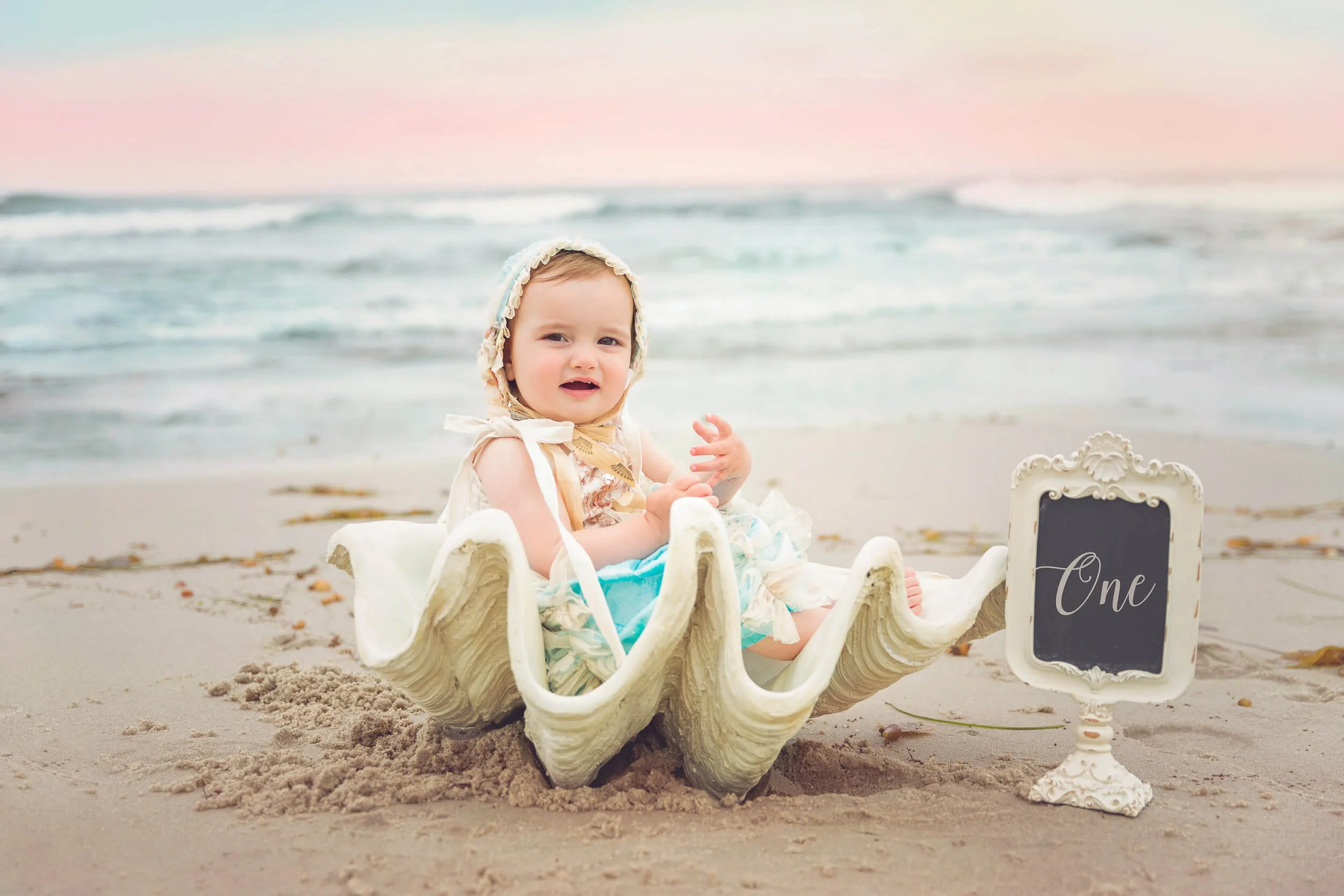 A baby sitting inside a big shell on the beach, holding her hands up, with a pastel sunset sky and ocean waves in the background, and a small framed sign that reads "One" next to her.