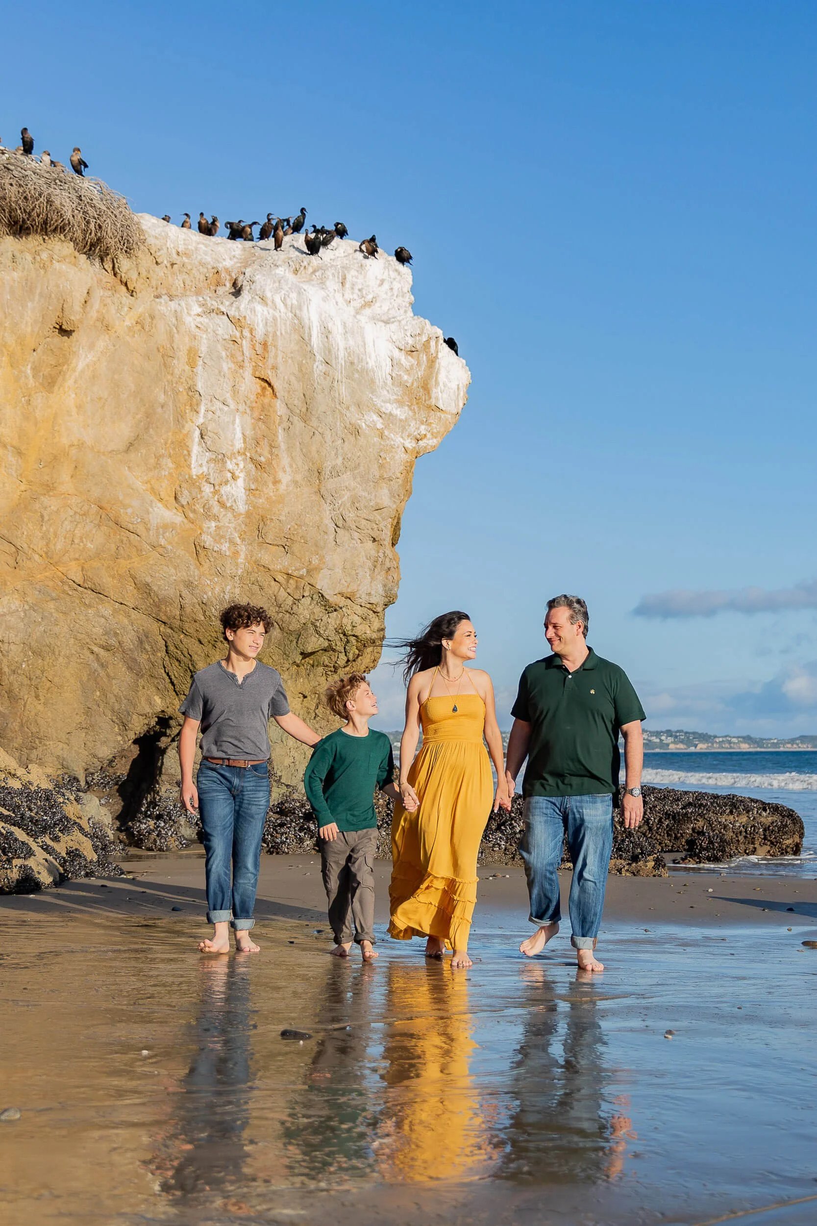 A family of four walking barefoot on the beach near a large cliff, with seagulls perched on top of the cliff and the ocean in the background on a clear day.
