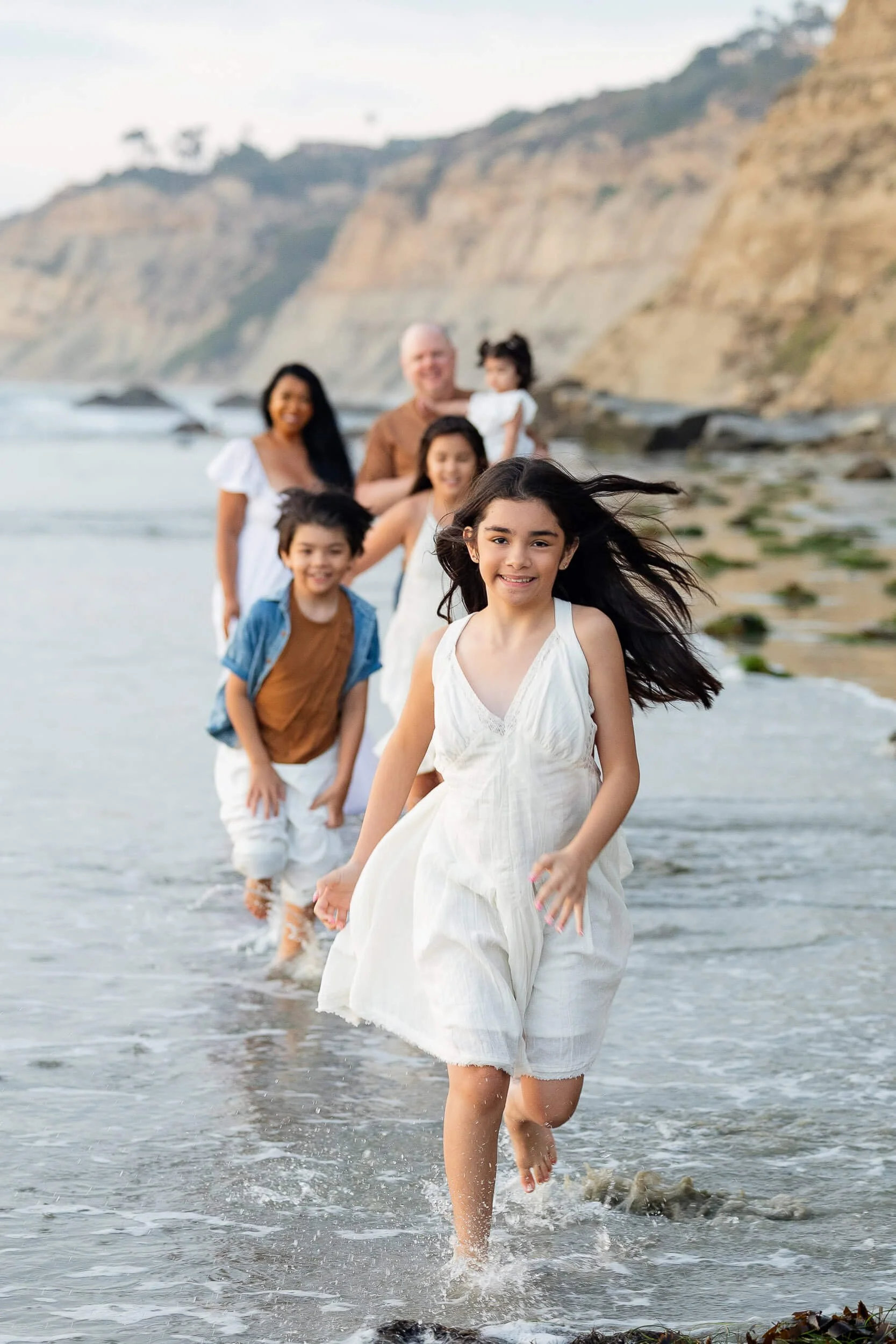 A family of seven running along the shoreline of a beach, with rocky cliffs in the background. The children are at the front, with a girl in a white dress leading, followed by other children and adults smiling.