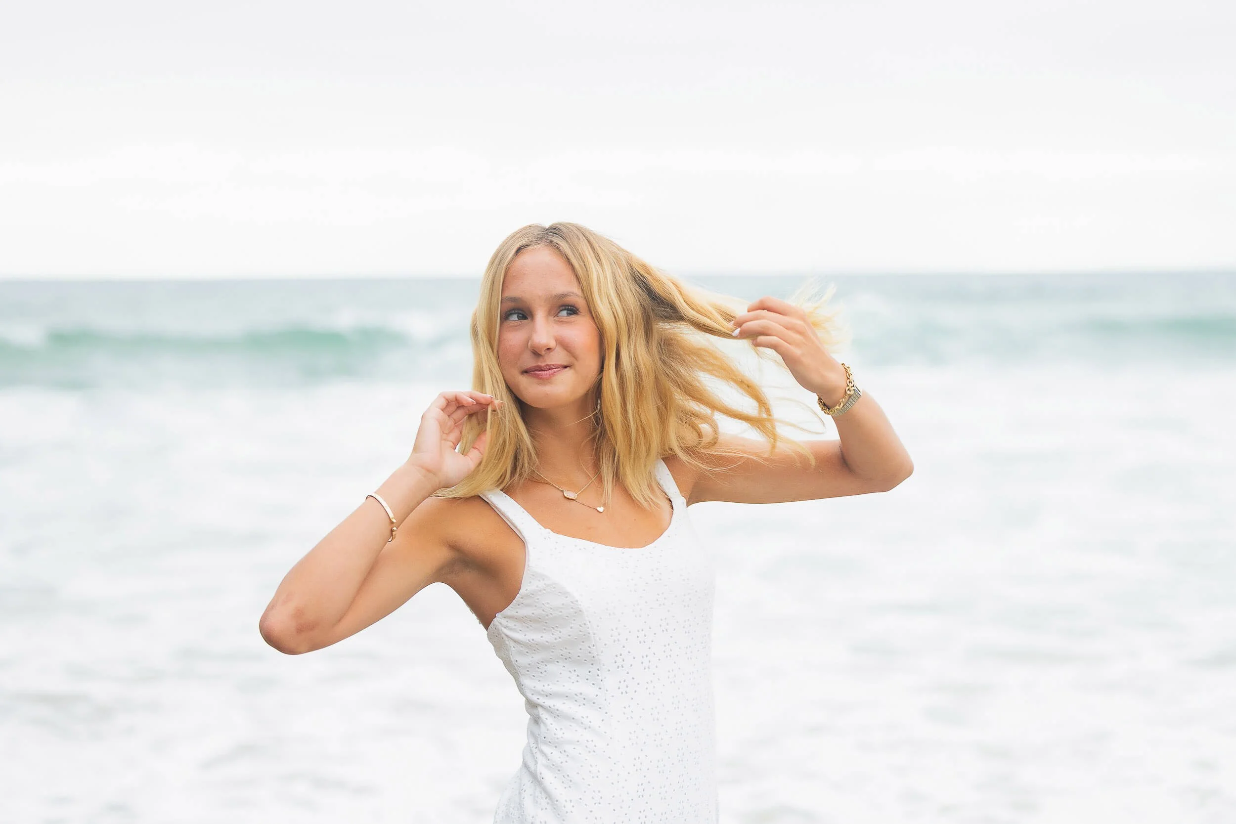 A young woman with blonde hair on a beach, wearing a white sleeveless dress, holding her hair, with the ocean in the background.
