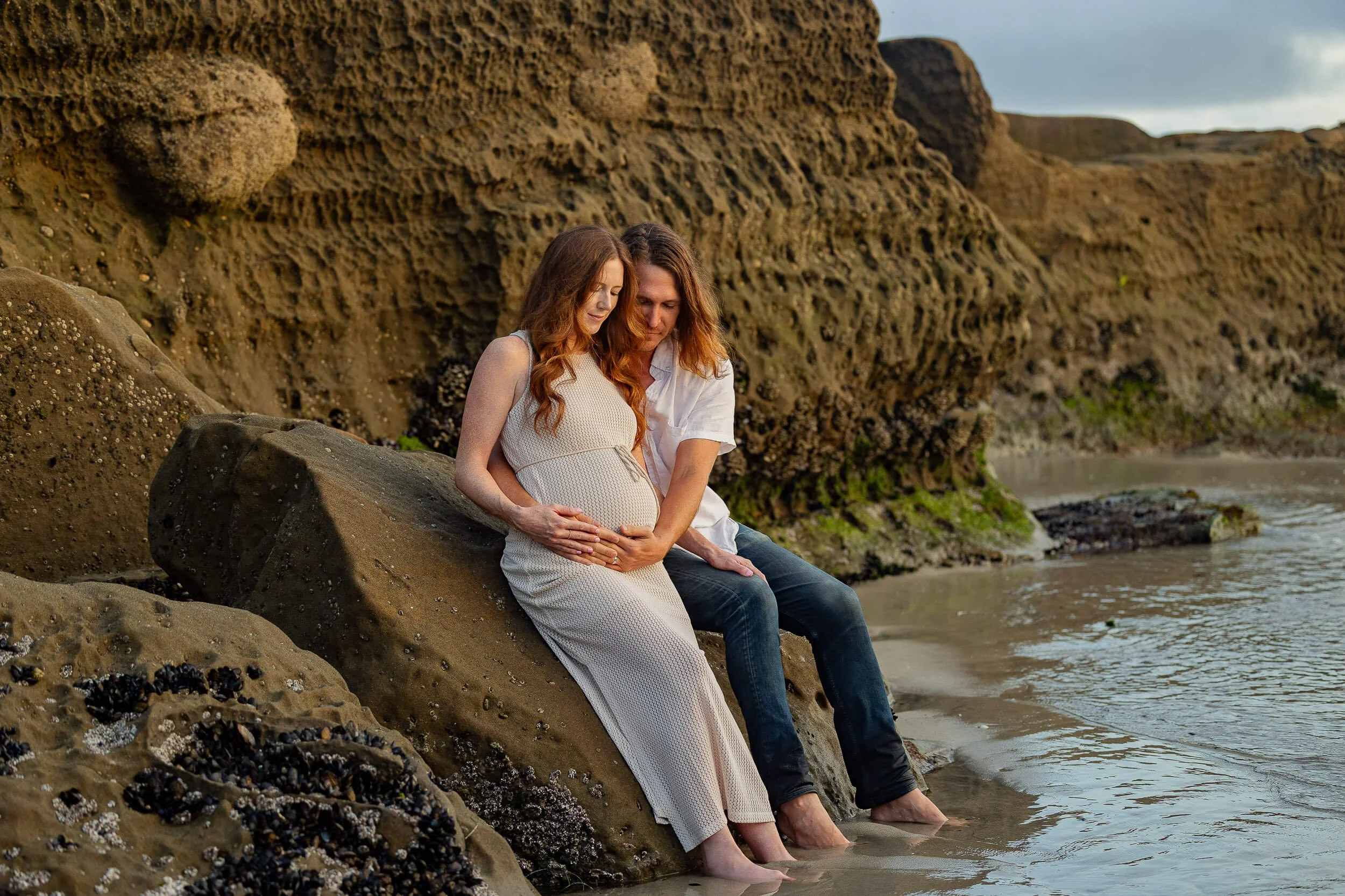A pregnant woman and a man sitting on a large rock by the beach, with rugged cliffs in the background, both with their feet in the water, looking down at her belly.