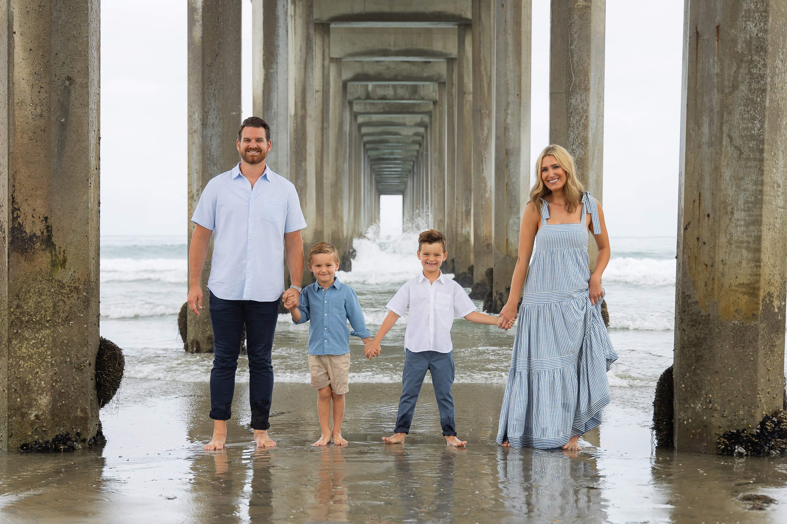A family of four standing on a beach under a pier holding hands and smiling, with ocean waves in the background.
