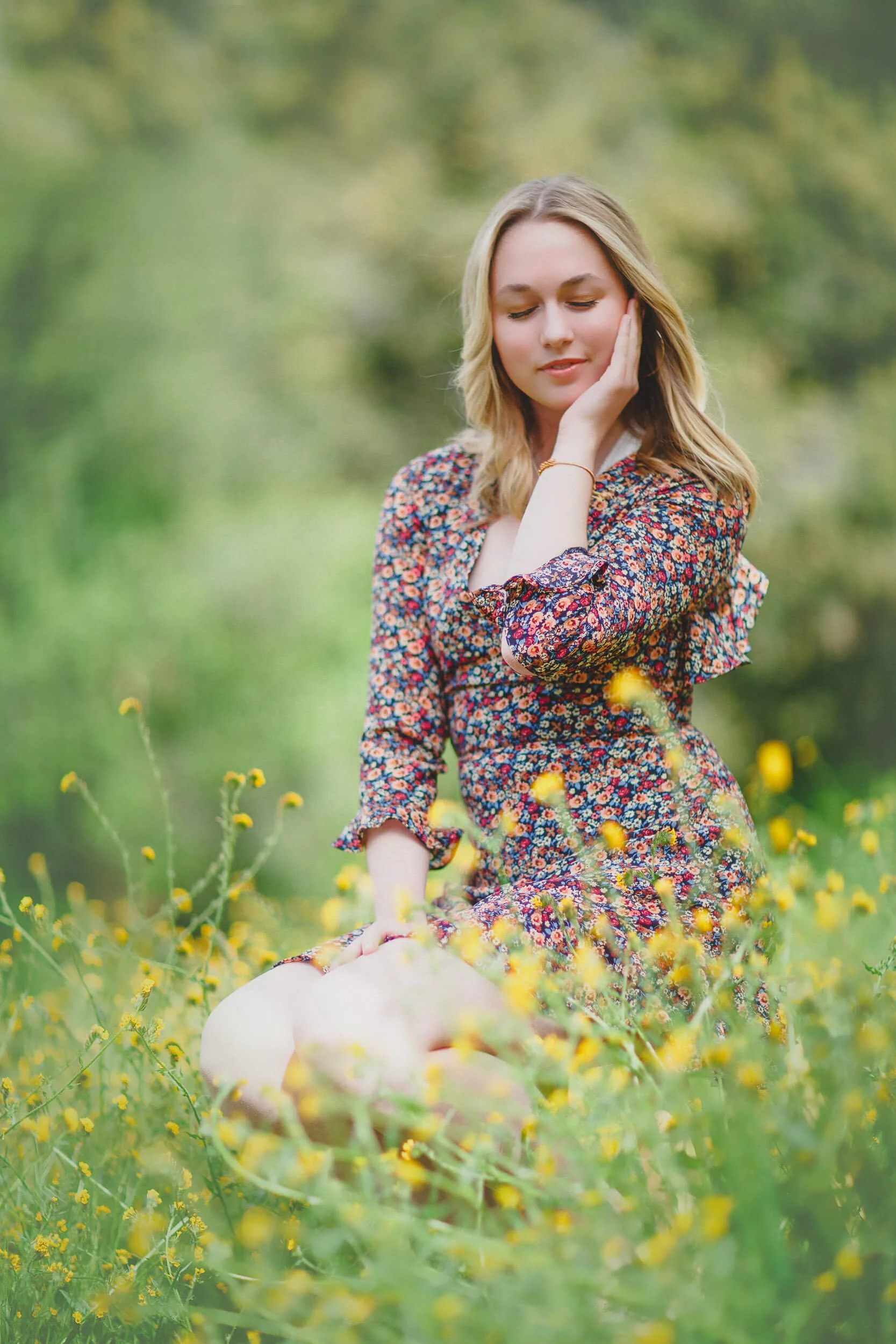 A young woman in a floral dress sitting in a field of yellow flowers, with her eyes closed and one hand touching her face.
