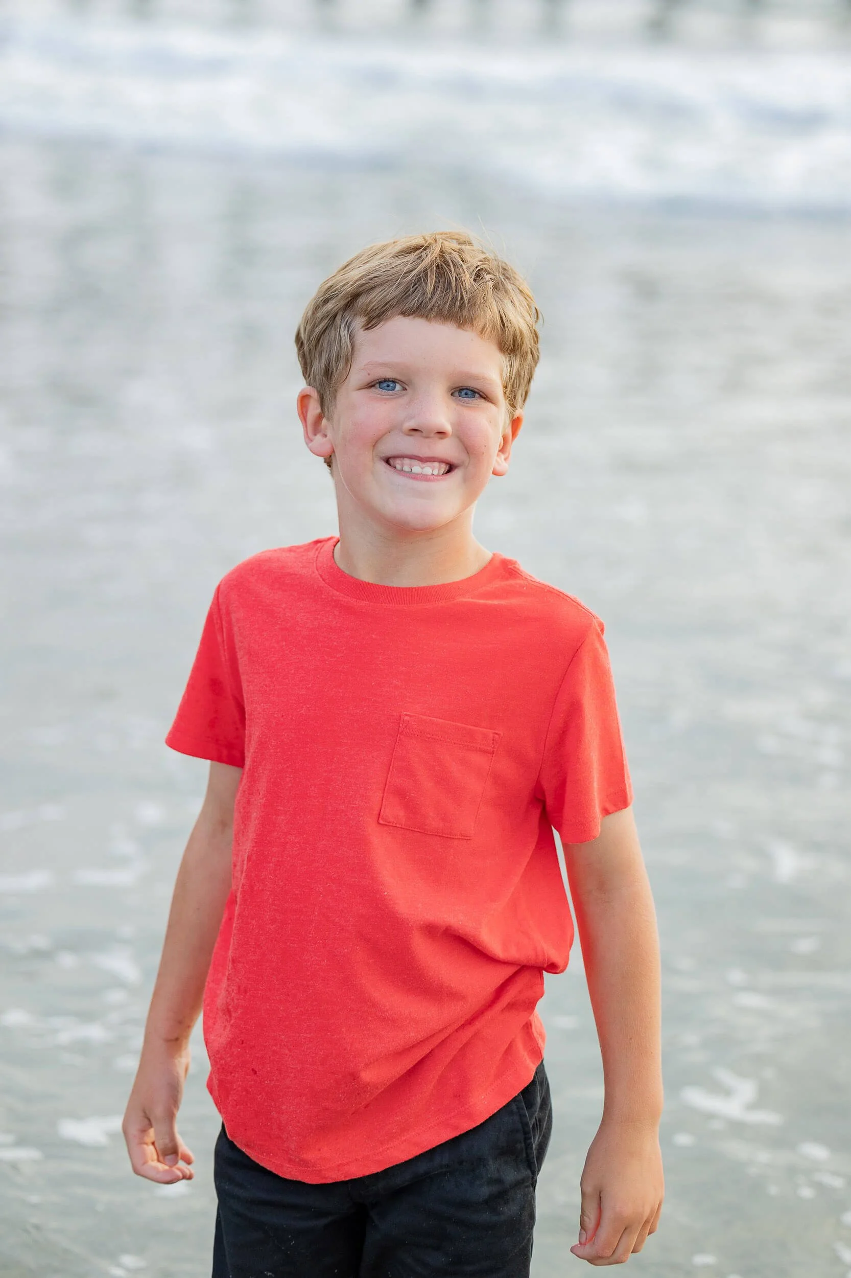 A smiling young boy standing in shallow water at the beach wearing a red t-shirt and black shorts.