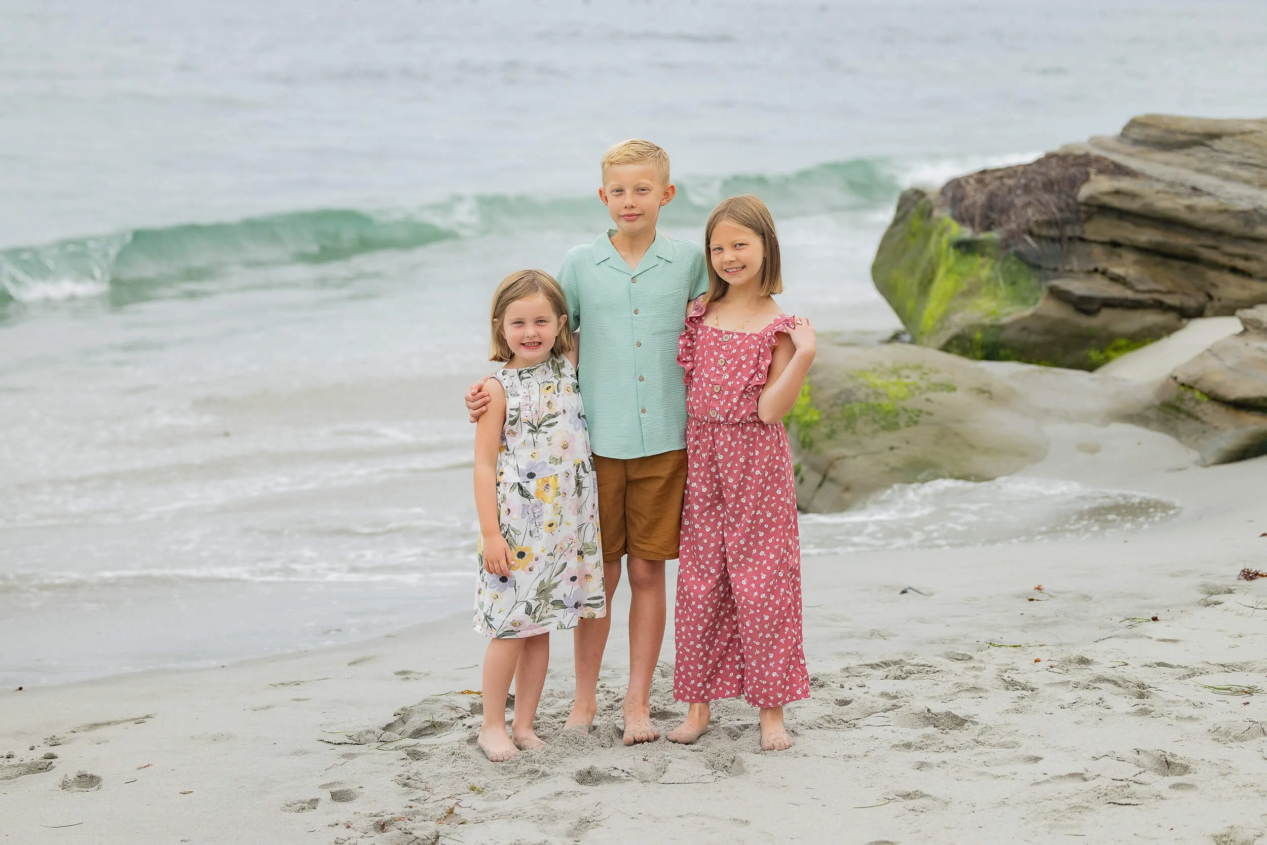 Three children, two girls and one boy, standing on the beach near rocks with waves in the background.