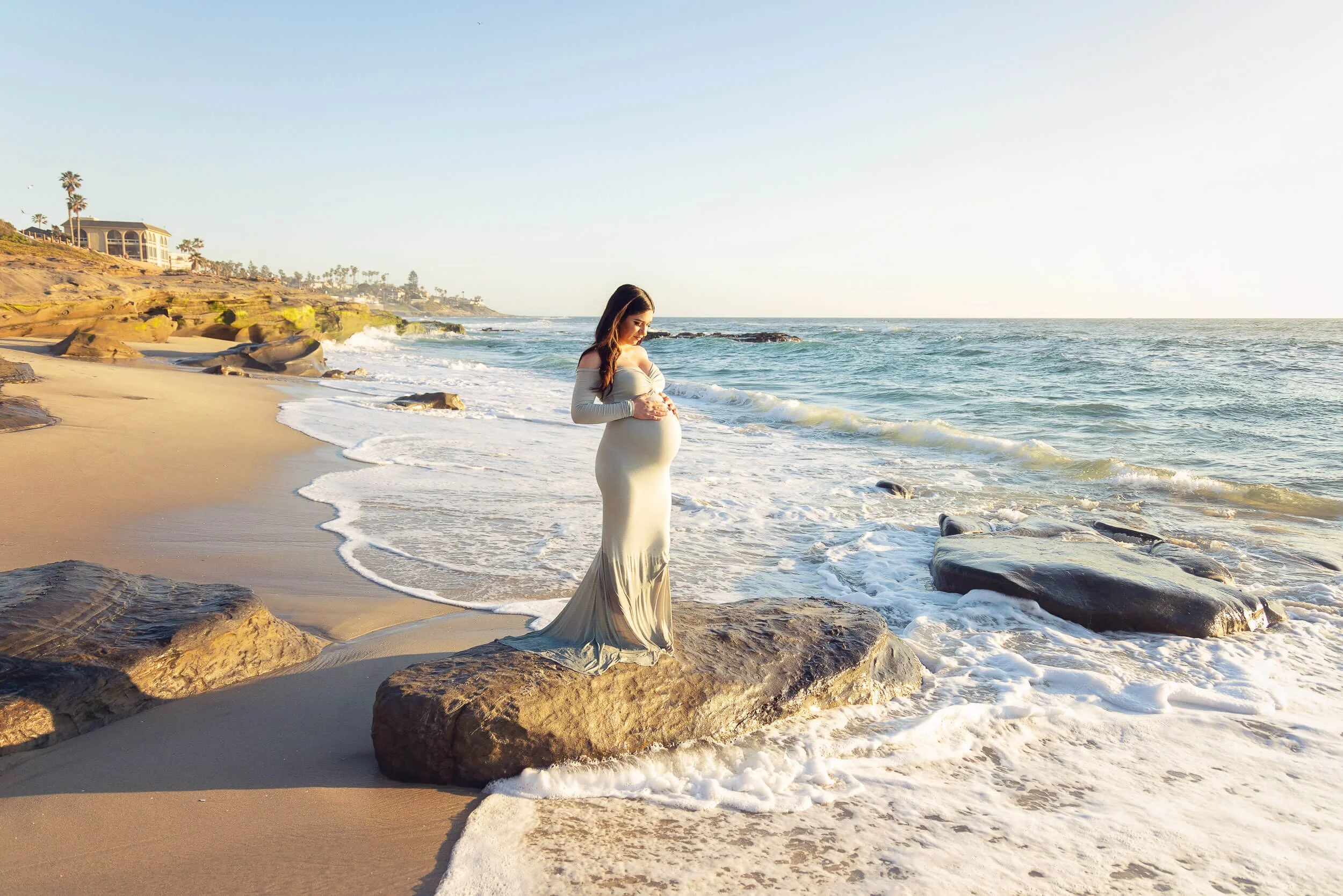 Pregnant woman in a long white dress standing on rocks by the beach, looking down at her belly with the ocean and shoreline in the background during sunset.