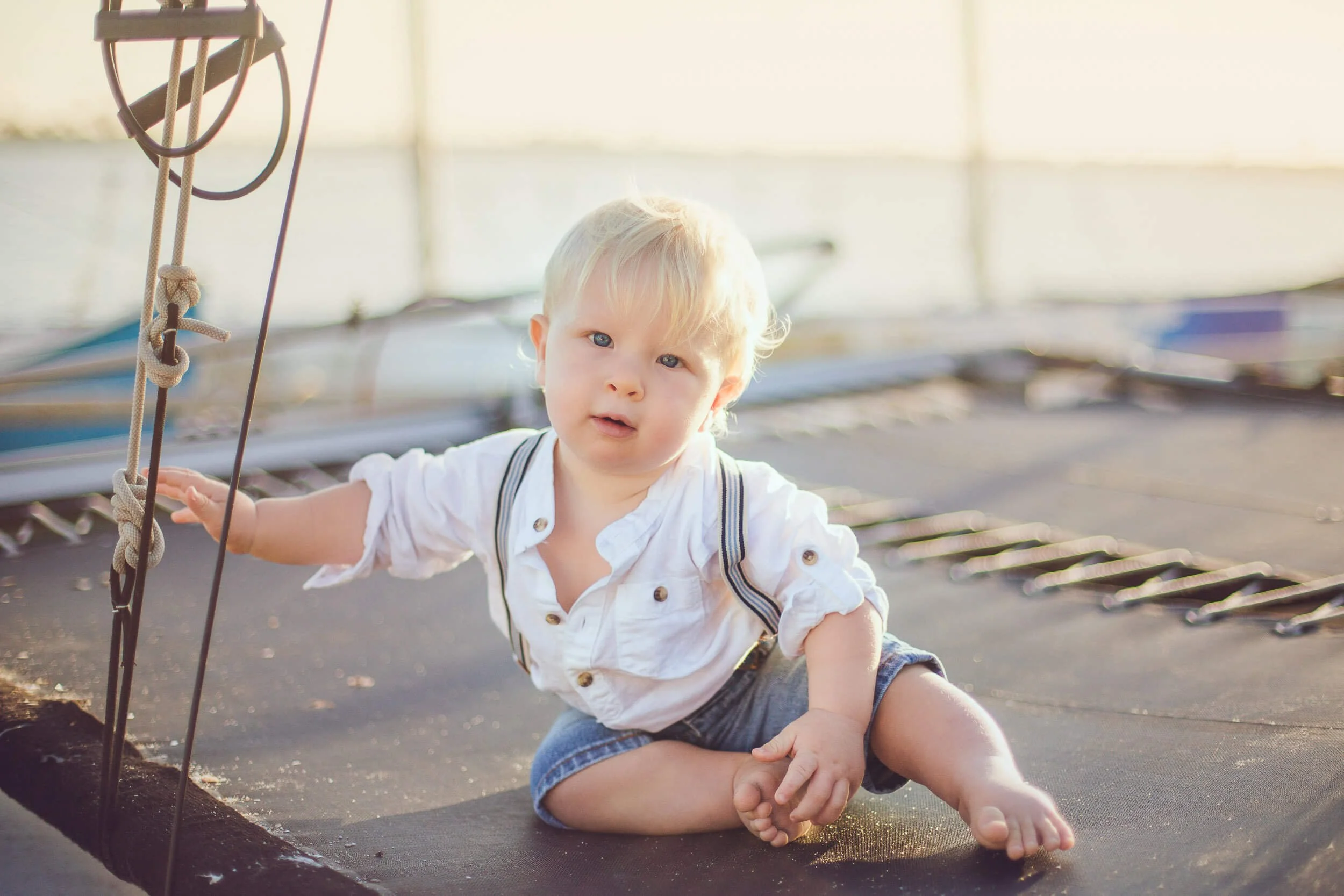 Young boy with blonde hair, sitting on a trampoline, wearing a white shirt with rolled-up sleeves and denim shorts, holding onto the trampoline's safety net.