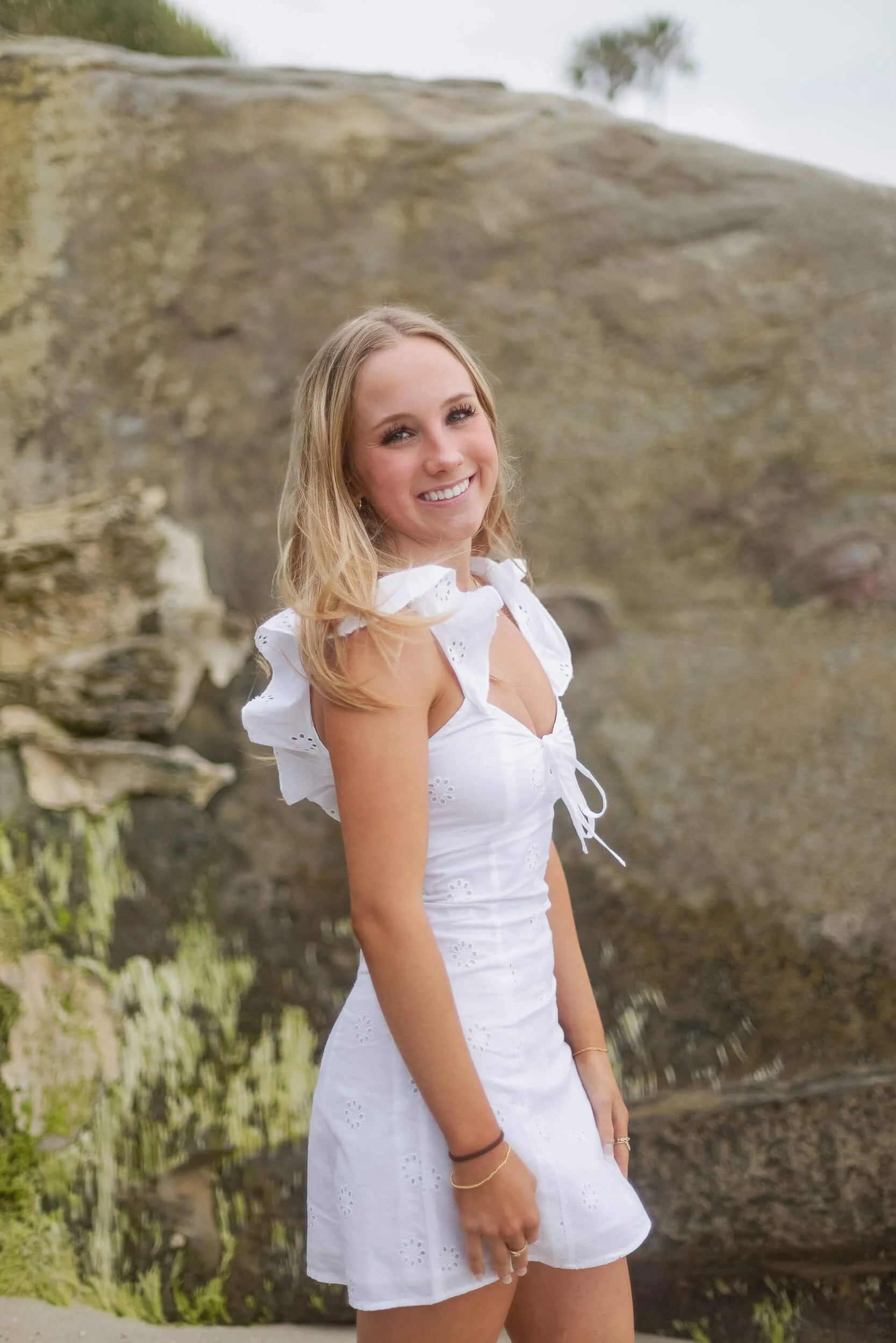 A young woman with blonde hair wearing a white eyelet dress with ruffled sleeves, smiling outdoors near a waterfall or rocky landscape.