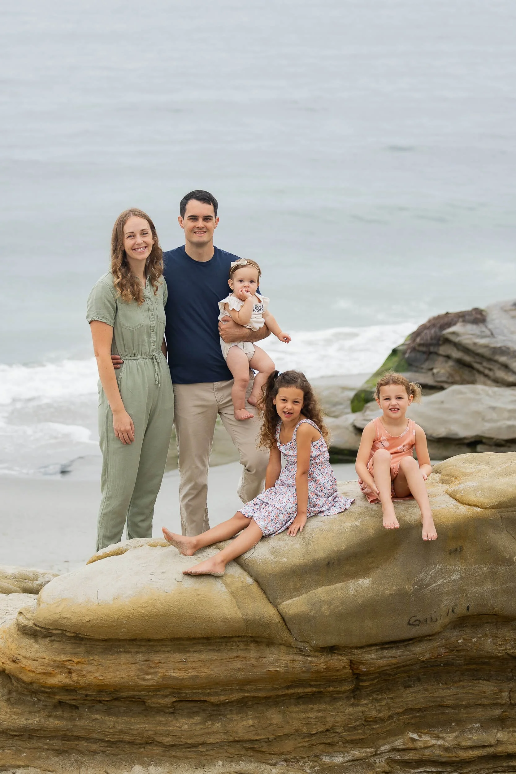 A family of five on rocks at the beach with the ocean in the background, including a woman, a man holding a baby girl, and two young girls sitting on a large rock.