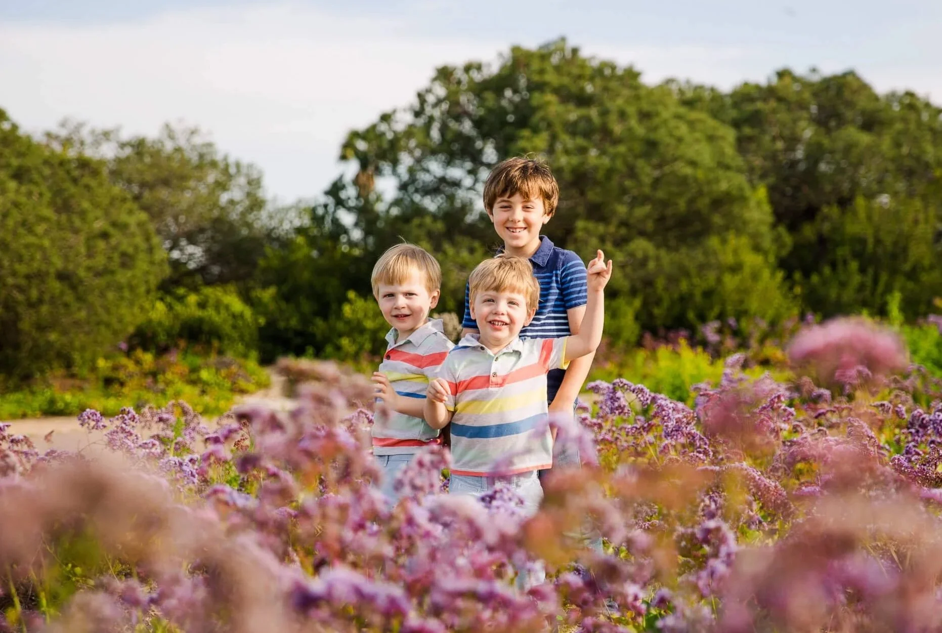 Three children, two younger boys and a teenage boy, standing among pink and purple flowers in a park with green trees in the background, smiling and enjoying a sunny day.