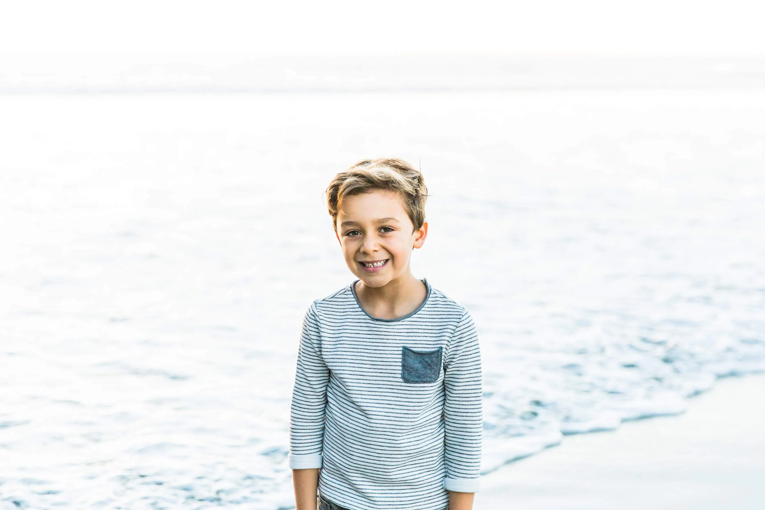 Smiling young boy with brown hair wearing a striped shirt standing in front of a water body at the beach