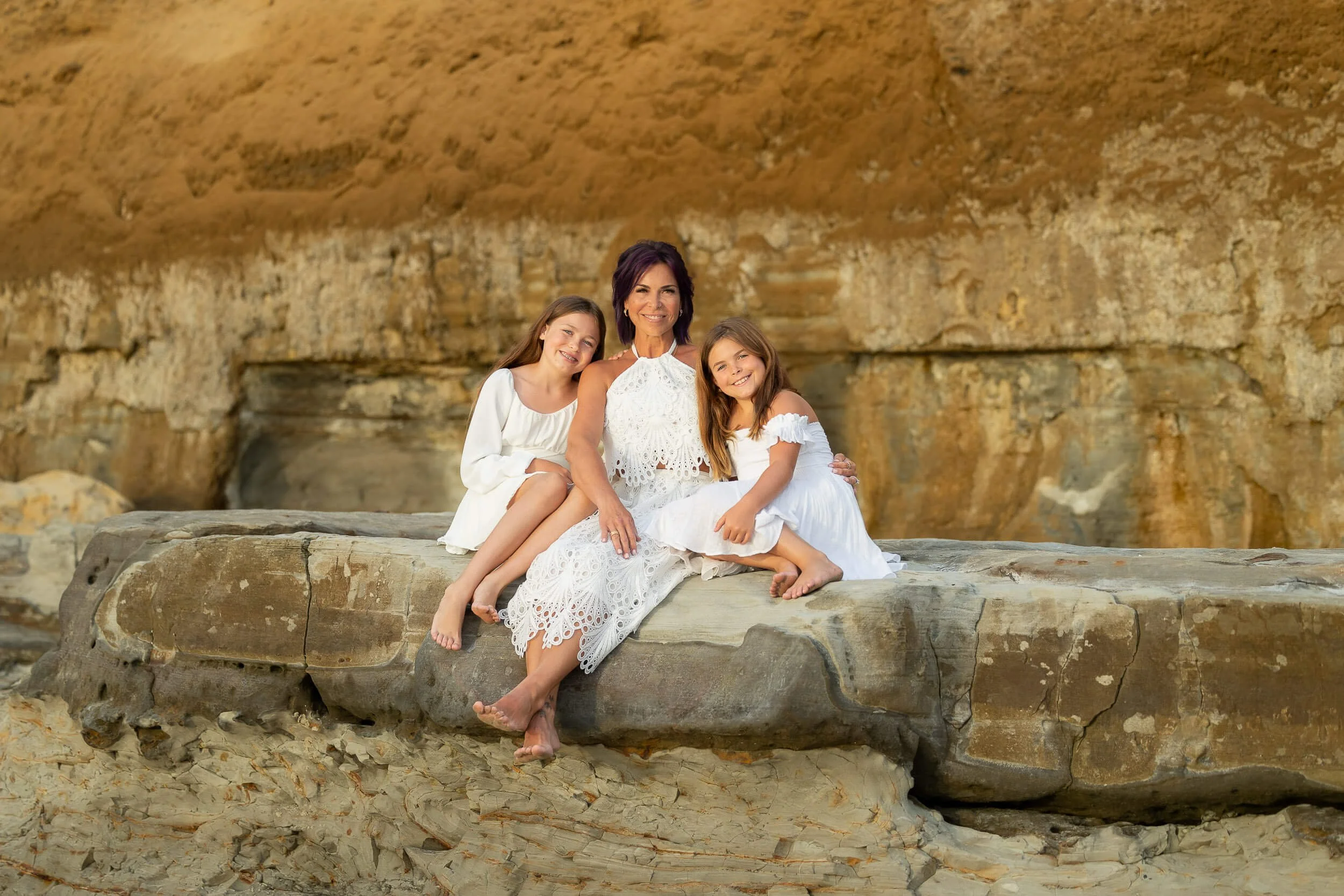 A woman and two young girls sitting on a large rock at the beach with a cliff in the background, all dressed in white and smiling.
