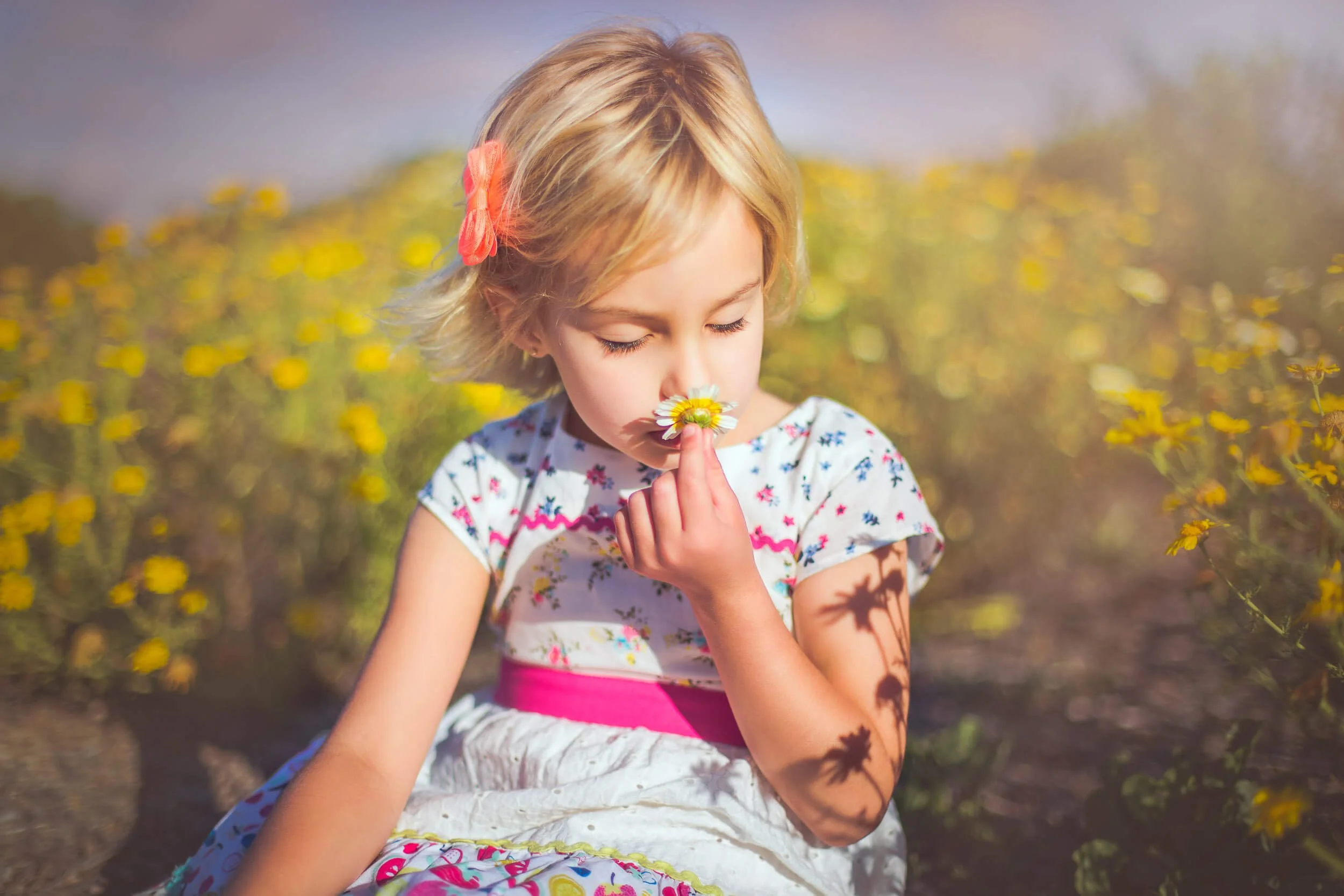 A young girl with blonde hair, wearing a floral dress with pink accents, sitting in a field of yellow flowers, smelling a daisy.