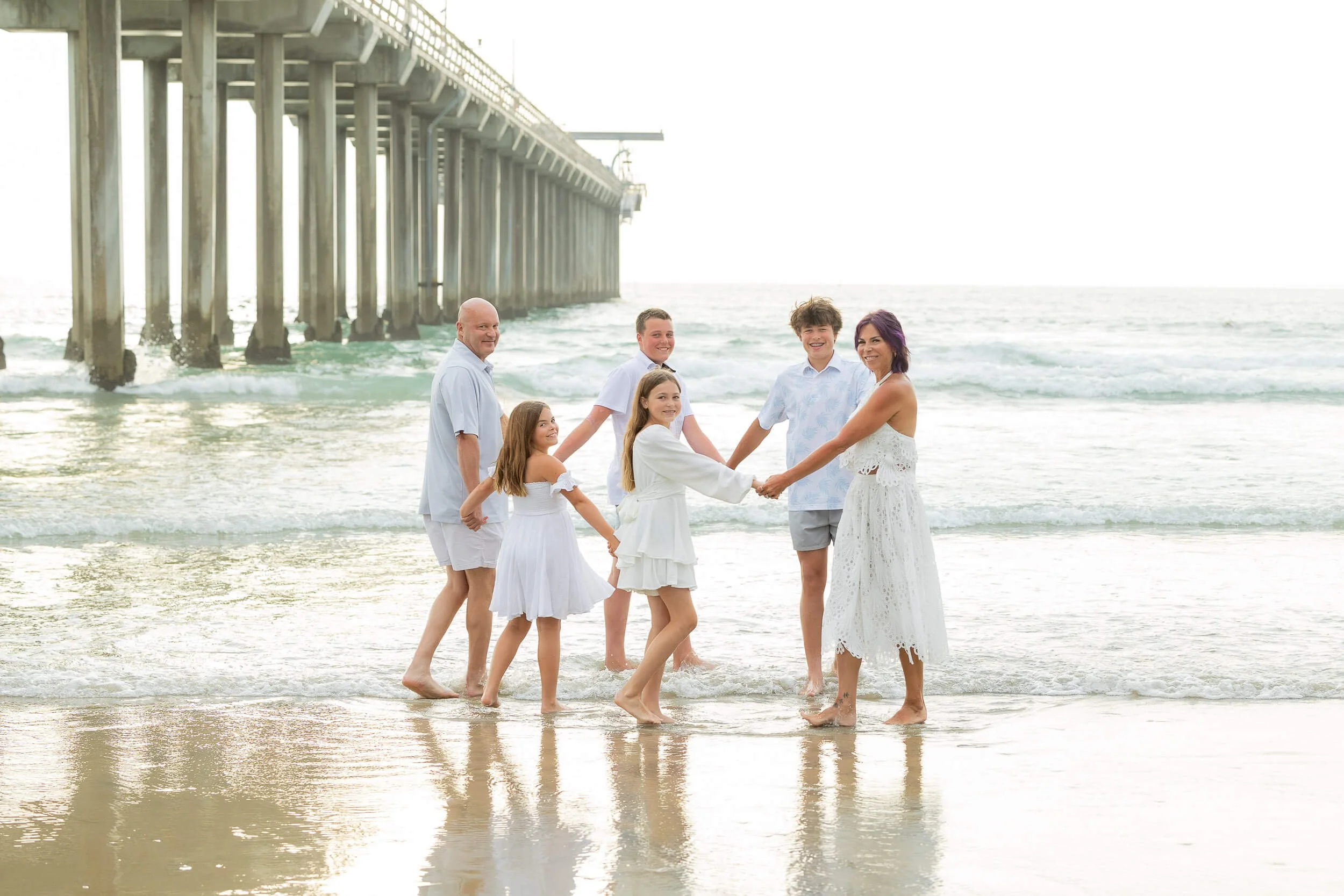 A family holding hands in a circle on the beach, near the ocean, with a pier in the background.
